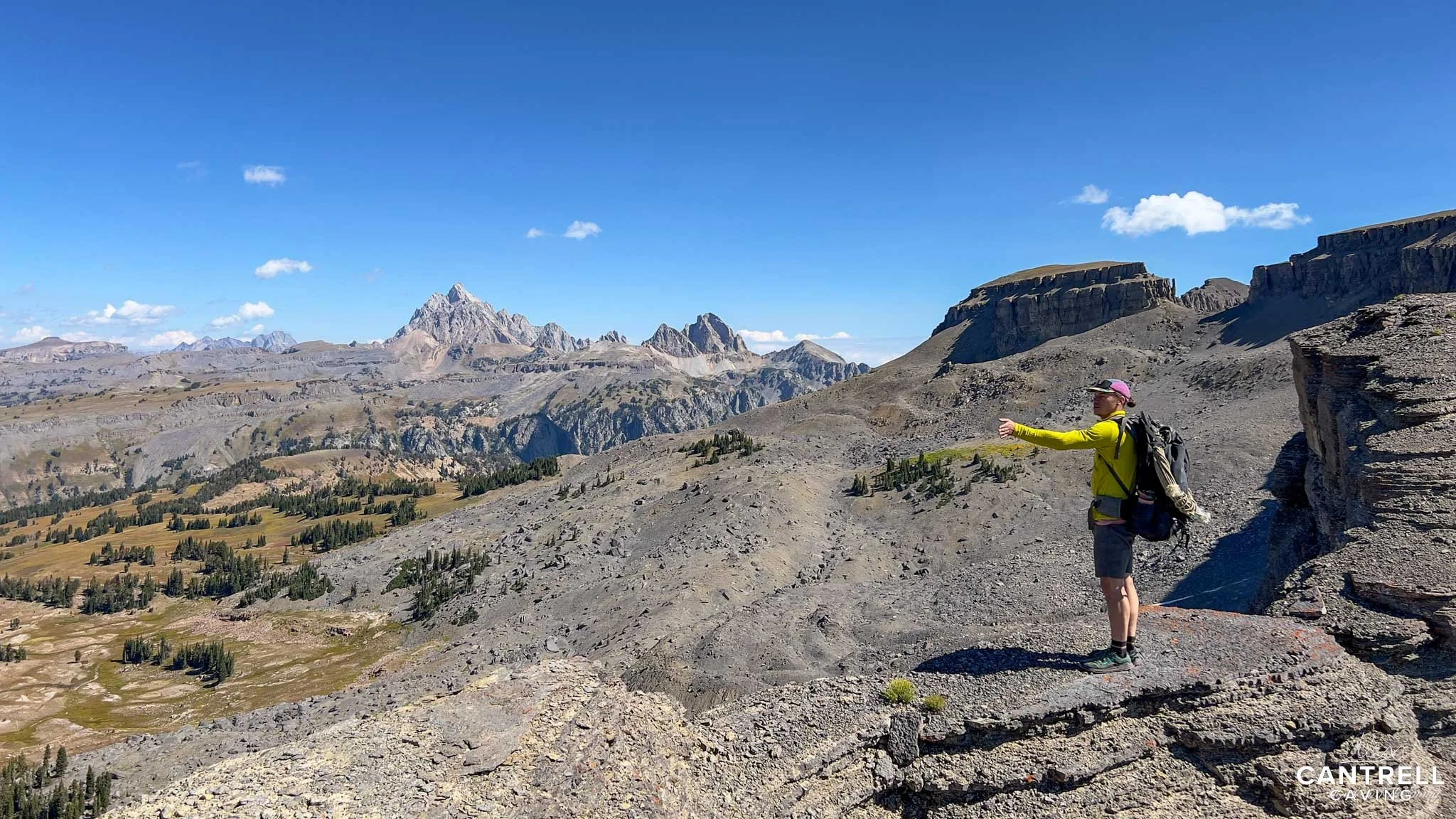 Hiker in yellow shirt with backpack pointing across a rocky plateau with mountainous landscape in the background under a clear blue sky.