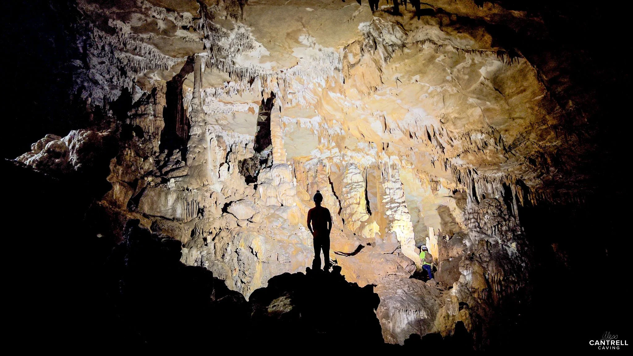 Silhouetted person standing in a dimly lit cave with stalactites and stalagmites.