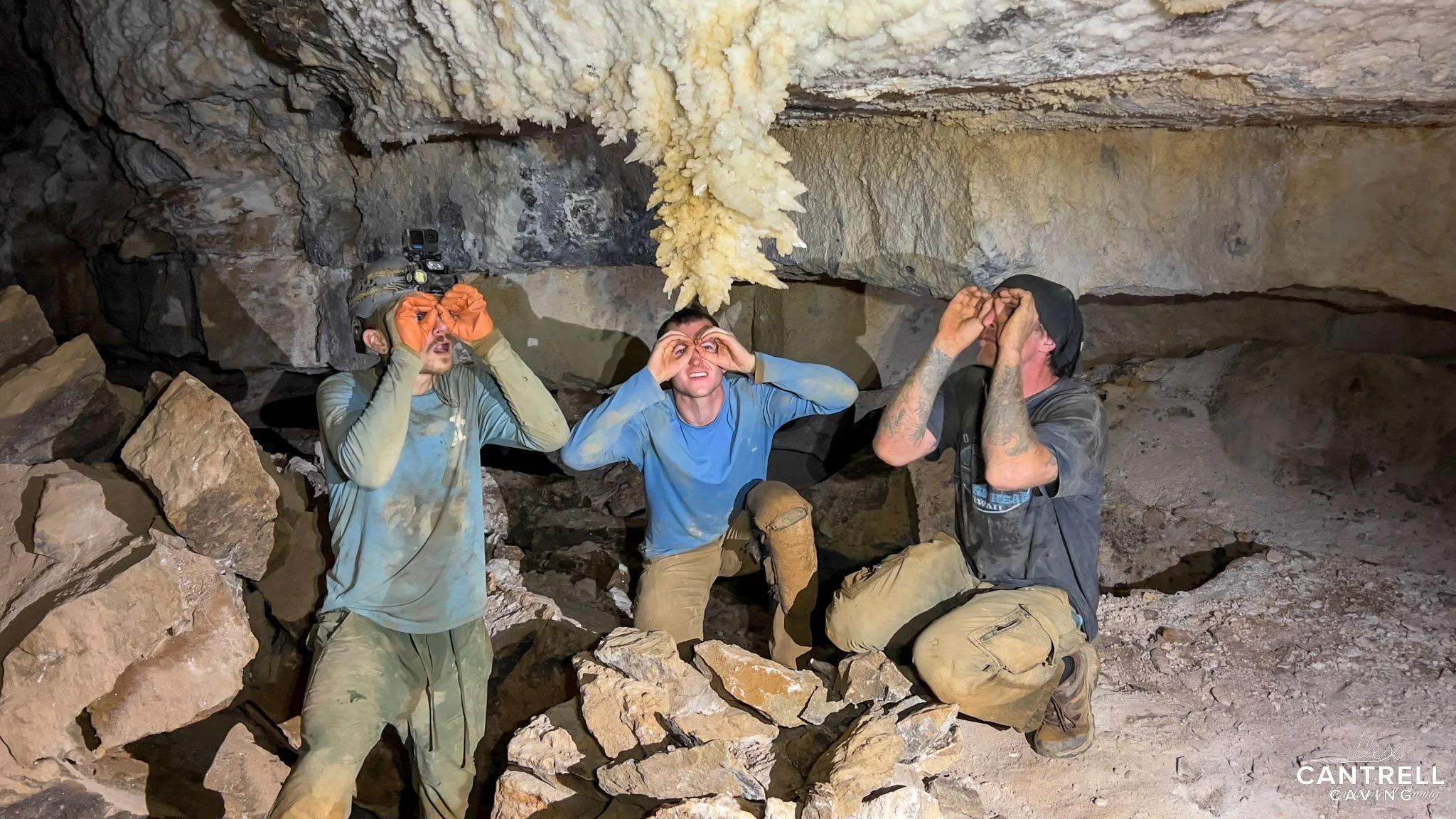Three people in a cave kneeling beneath a stalactite formation, wearing caving gear and making binocular shapes with their hands.