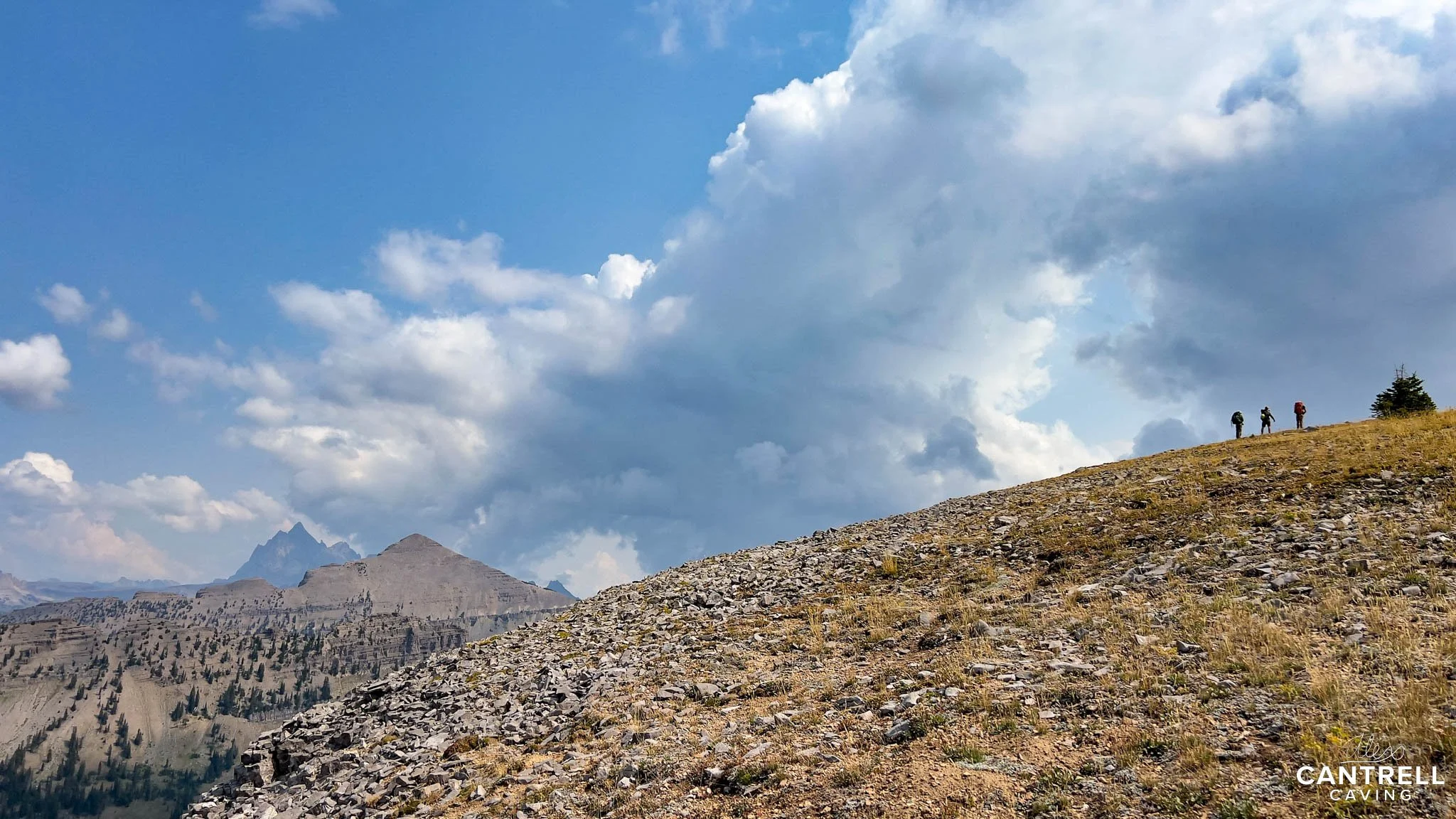 Hikers on a rocky and grassy hill with mountains and cloudy sky in the background.