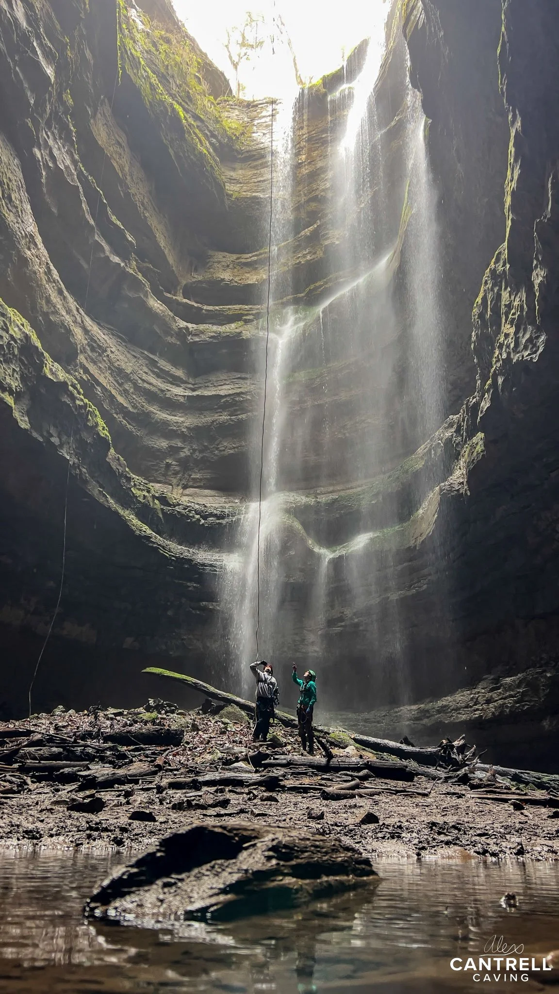 Cave entry with two people exploring; waterfall cascading down from above, natural light illuminating the scene.