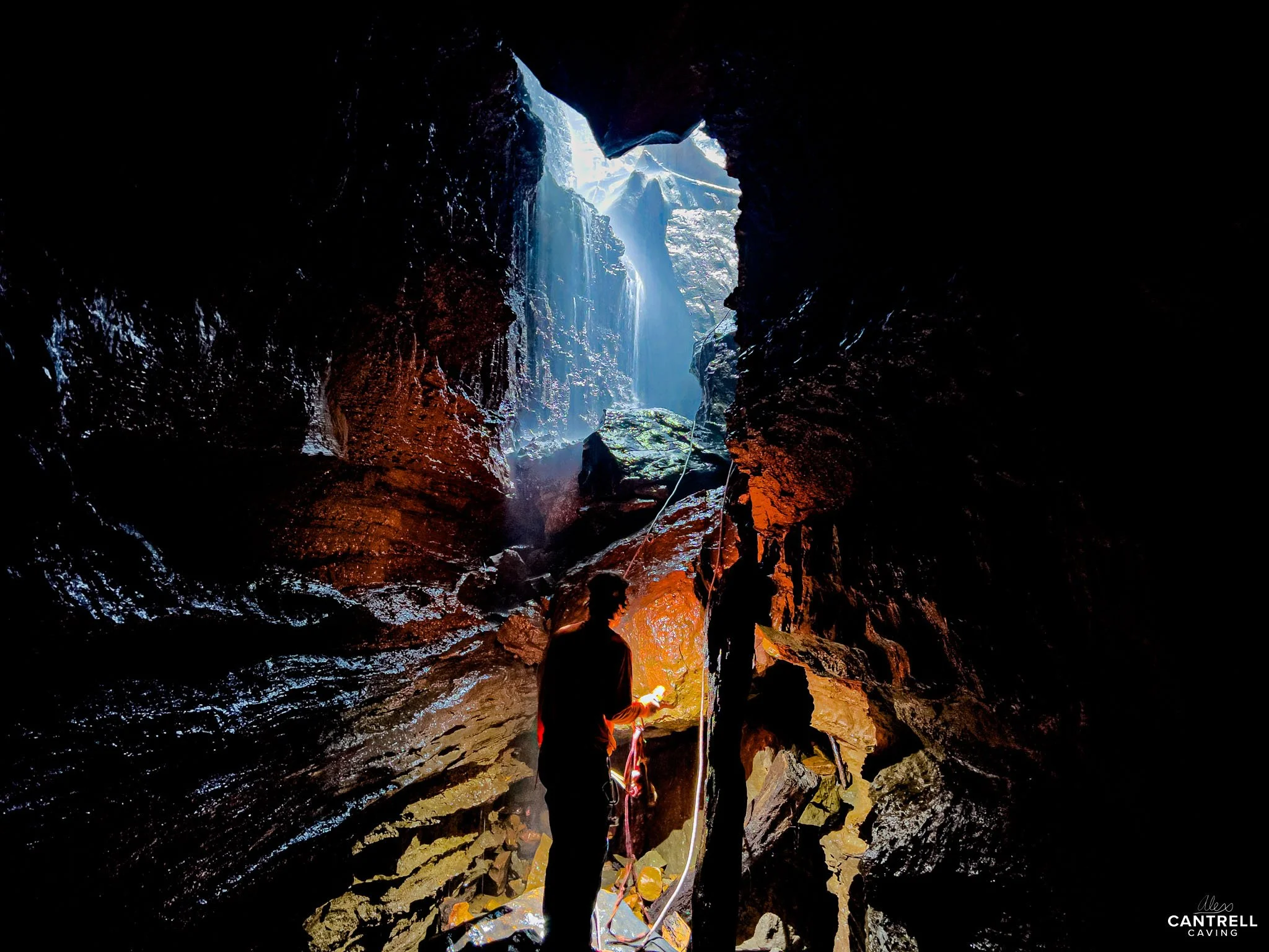 Person exploring a dark cave lit by natural light from an opening above, with wet and rocky walls.