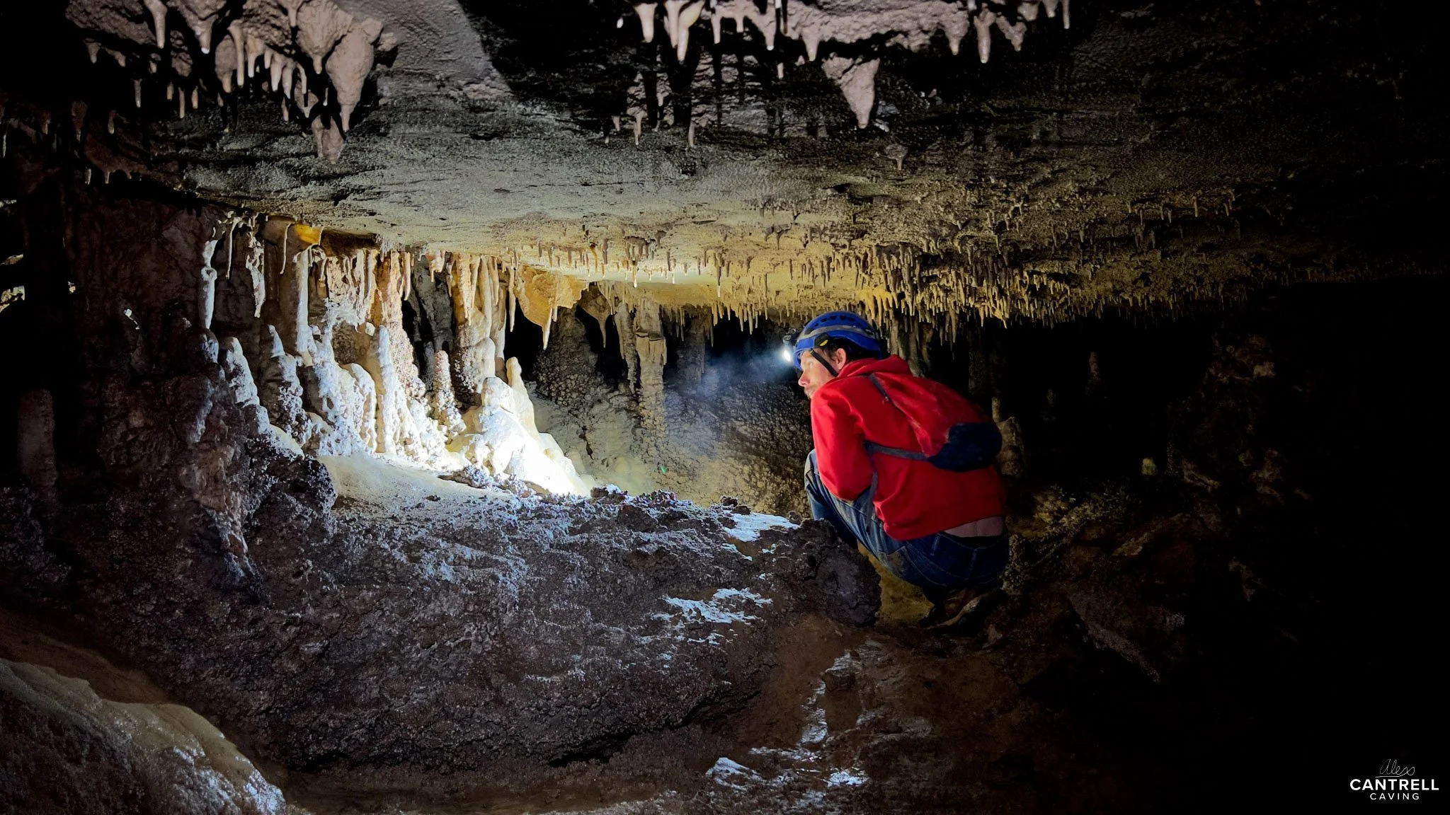 Person in red jacket and helmet exploring a cave with stalactites and stalagmites, using a headlamp for illumination.