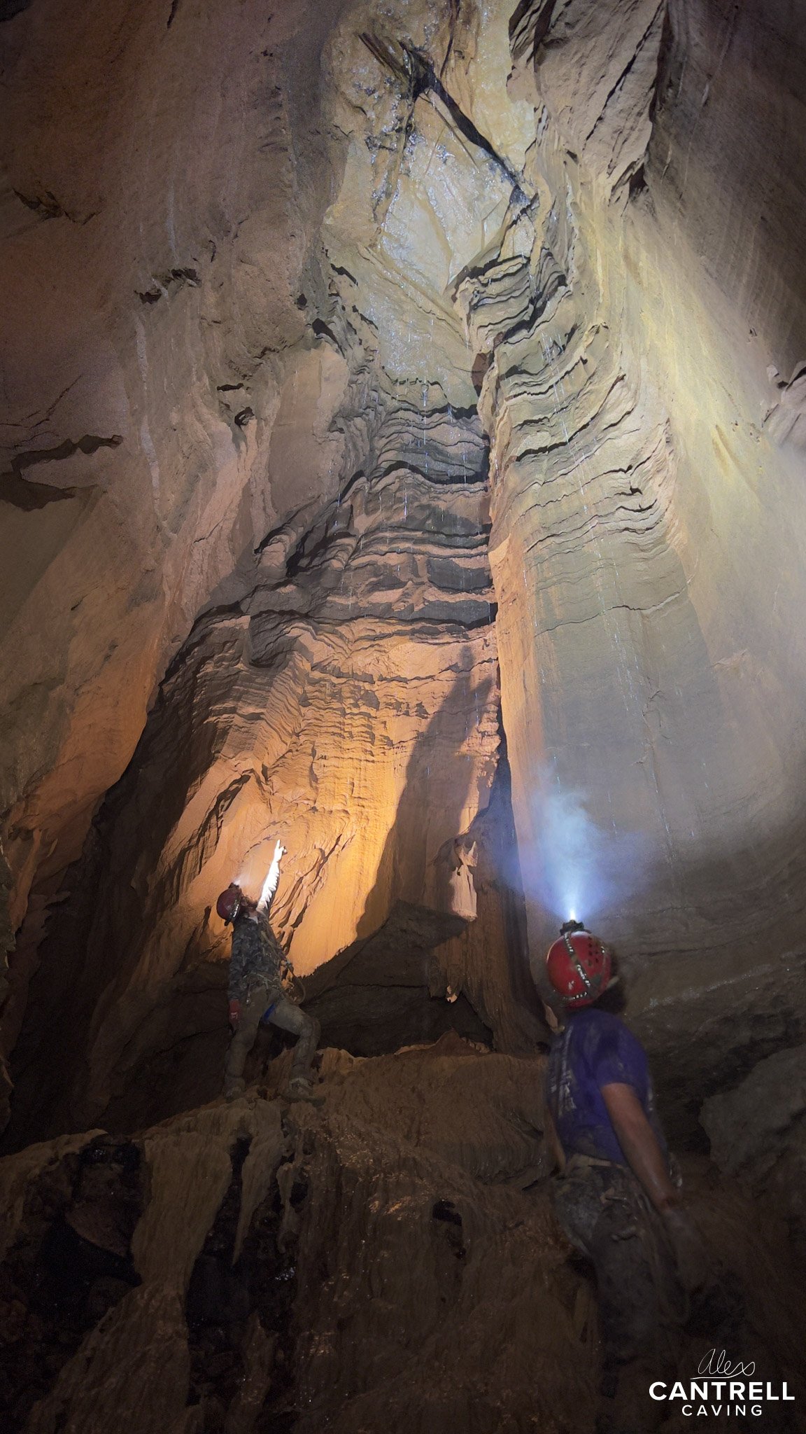 Two cavers exploring a large underground cave with layered rock formations, one pointing upward and both wearing helmets with lights.