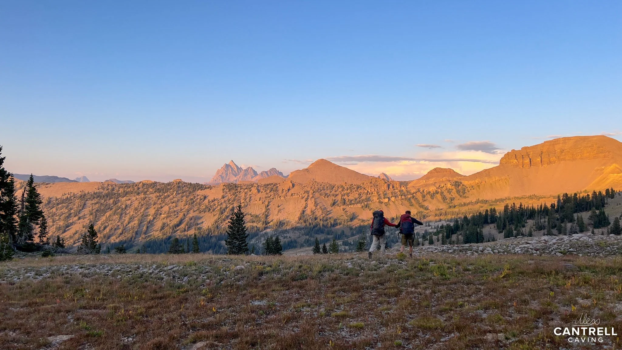 Two hikers with backpacks walking across a grassy meadow with distant mountains under a clear blue sky during sunset.