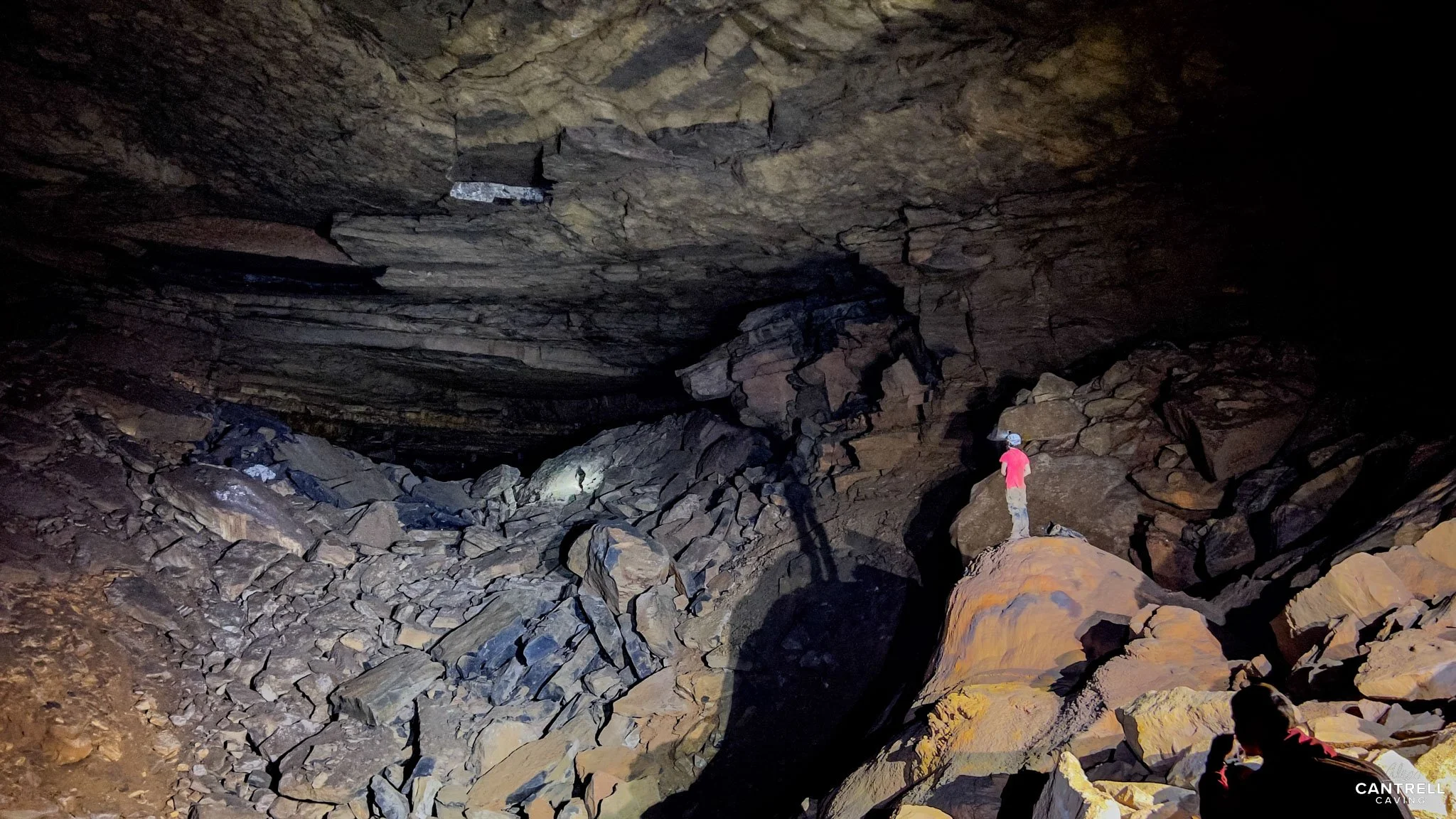 Person wearing a helmet standing inside a large cave with rocky terrain and dim lighting.
