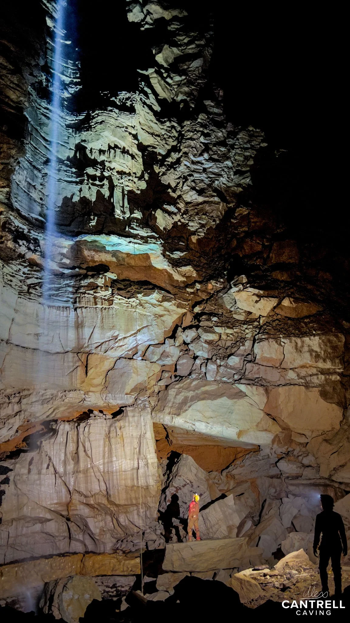 A large cave interior with rugged rock formations, illuminated by natural light and a beam of sunlight. A person in caving gear stands near the rock formations, showcasing the scale and depth of the cave. The environment is dimly lit, emphasizing the