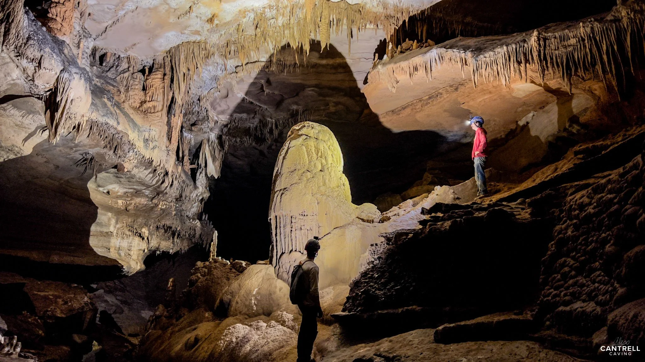 Two people exploring a cave with stalactites and stalagmites, one wearing a helmet with a headlamp, another standing on rocky ground. The cave features dramatic lighting and rock formations.