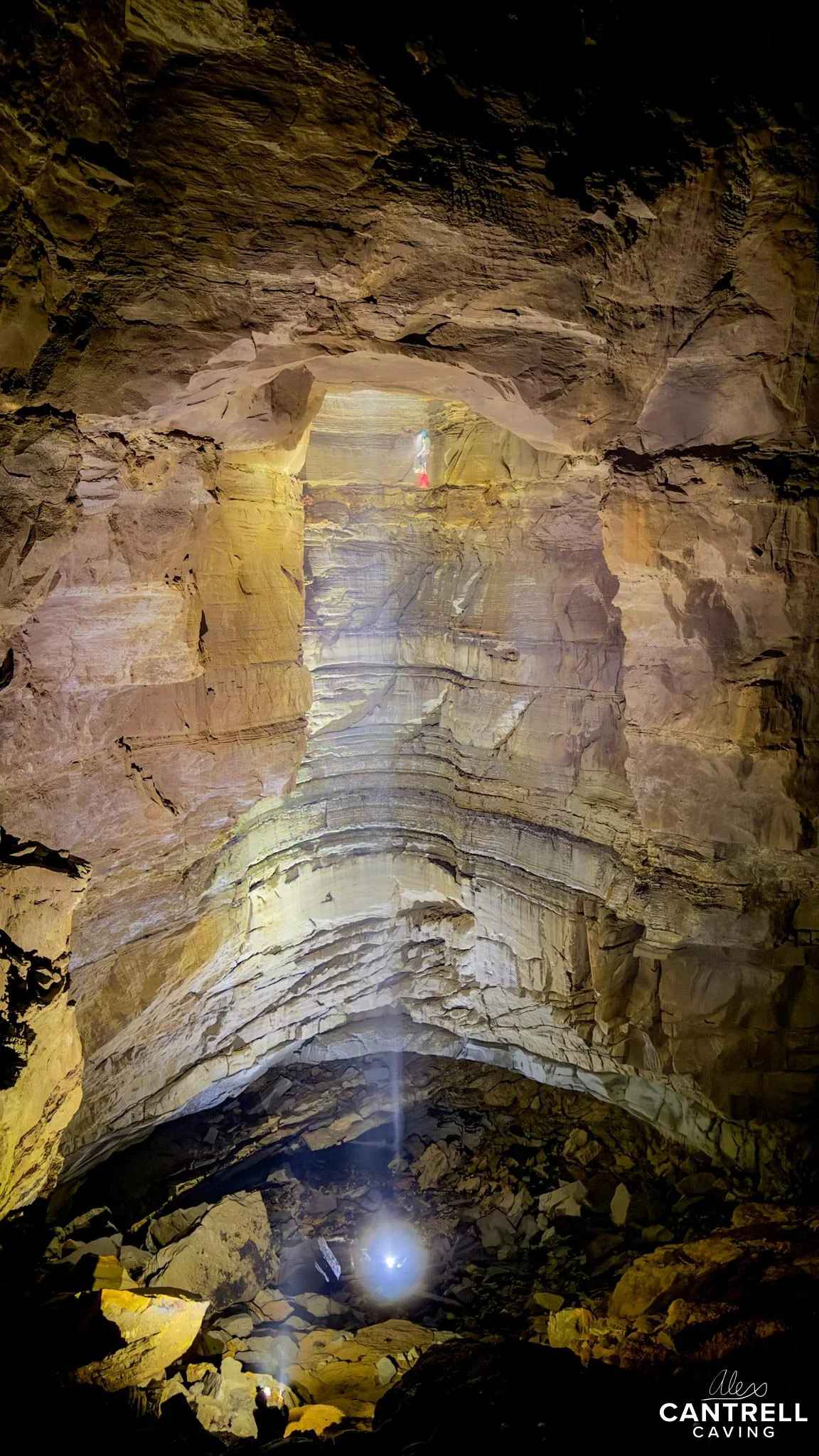 Person exploring a large, illuminated cave with rock formations and a bright light source at the bottom.