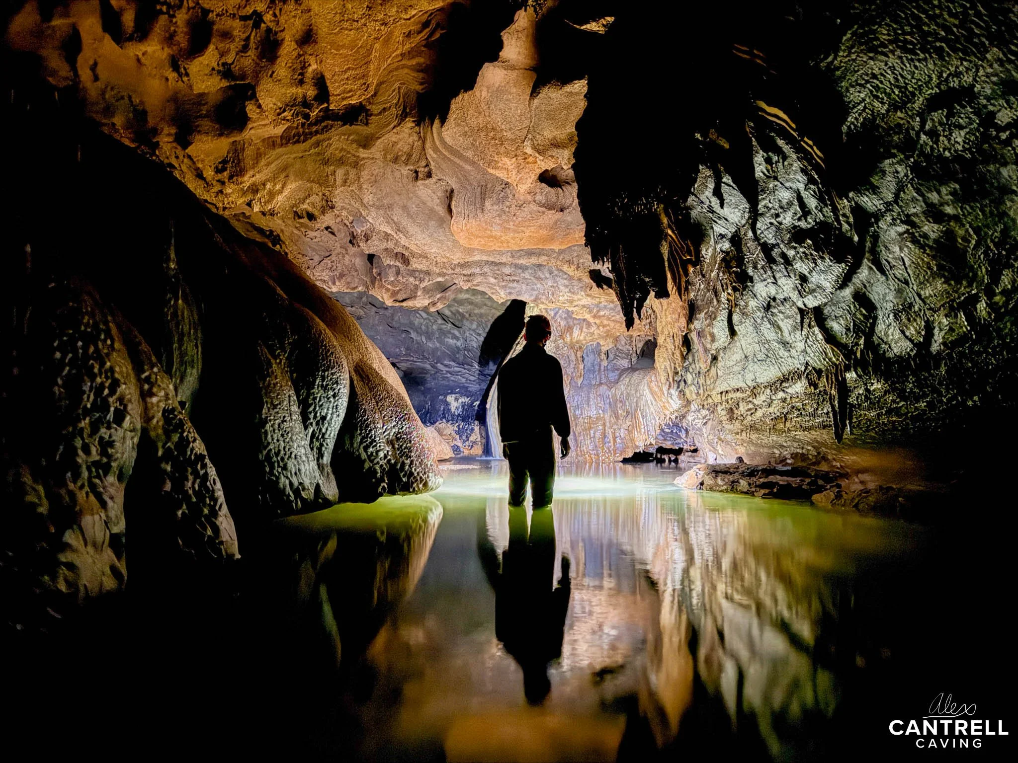 Person standing in a dimly lit cave with reflective water on the floor, surrounded by stalactites and rugged rock formations.