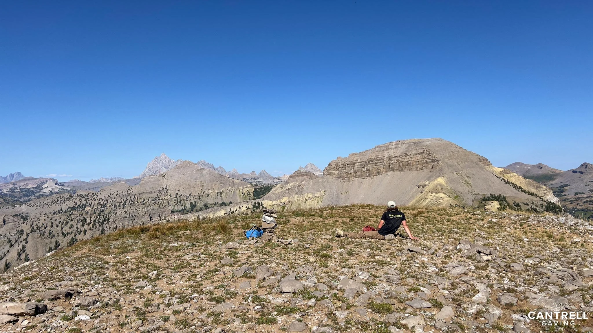 Person sitting on rocky mountain landscape with distant mountain peaks under clear blue sky.