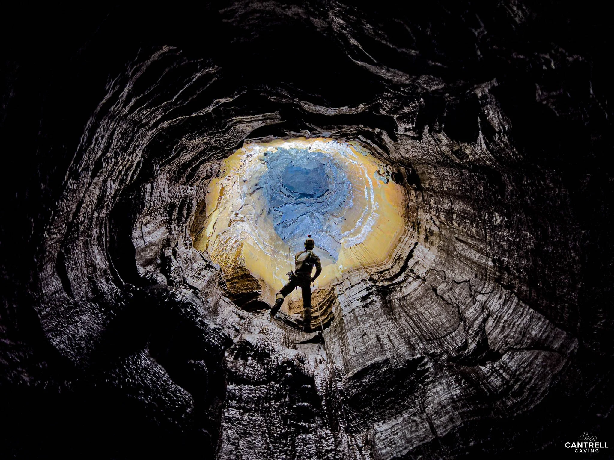 Person standing inside a cave, looking up at a circular opening with sunlight illuminating the rock formations.