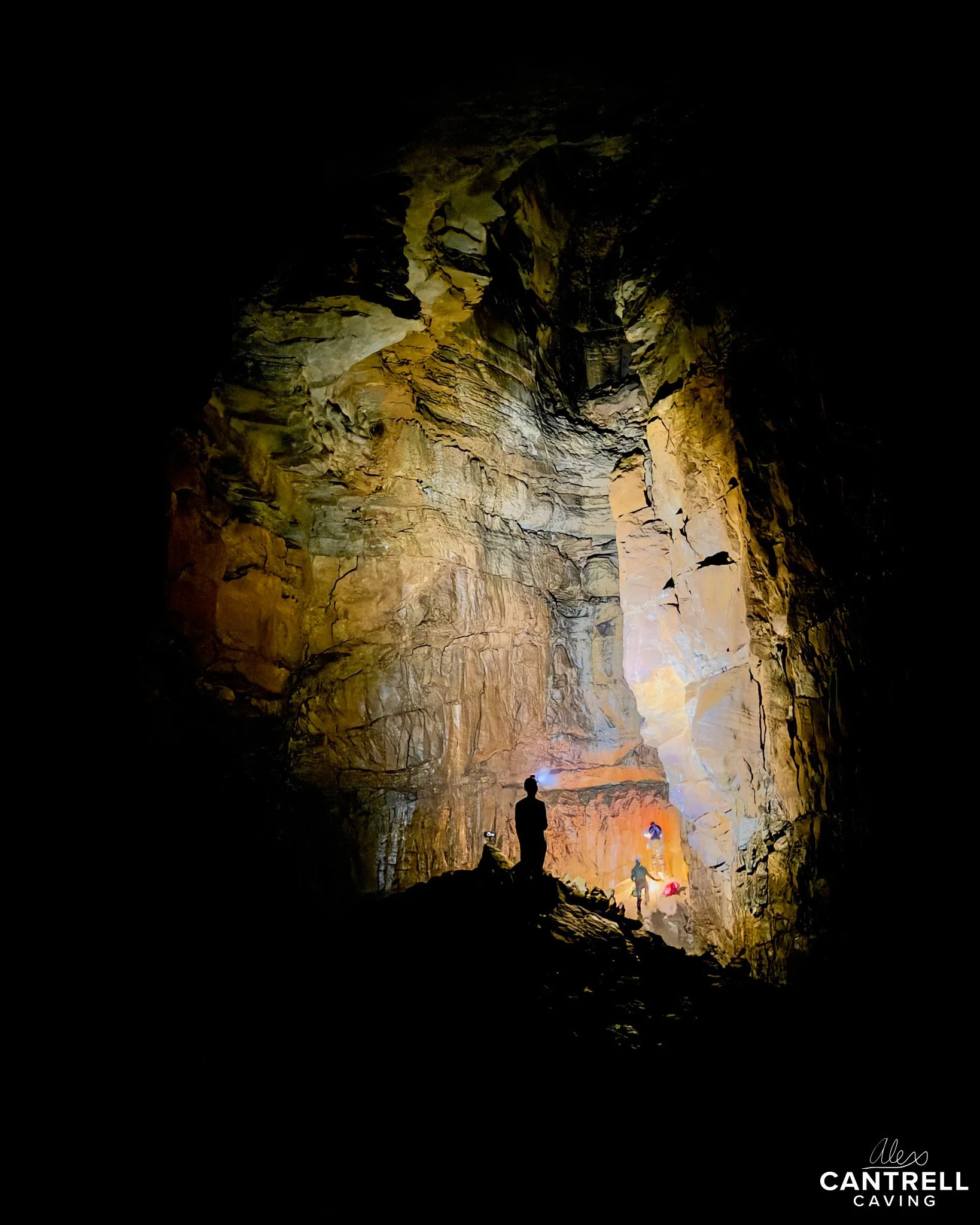 Cave exploration scene with two silhouetted figures, illuminated rock formations, and colorful light reflections on cavern walls. "Cantrell Caving" logo is visible.