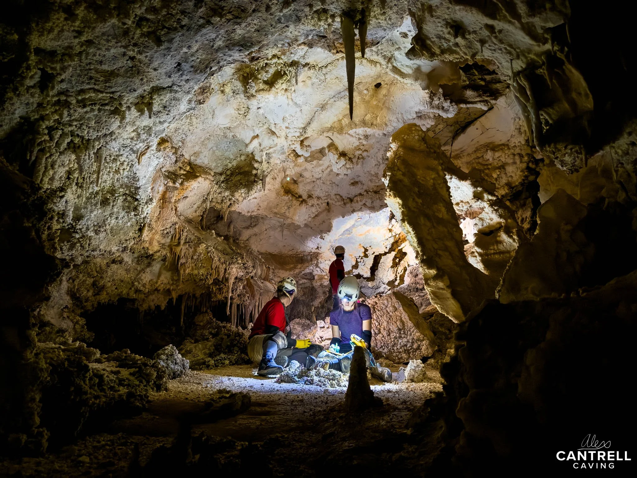 Three people wearing helmets exploring a cave; illuminated rocky interior with stalactites and stalagmites.