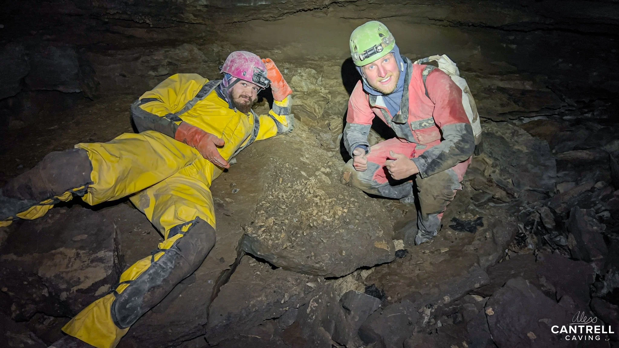 Two people in caving gear inside a cave, wearing helmets with headlamps, one in a yellow suit reclining on the ground, the other kneeling in a red suit.