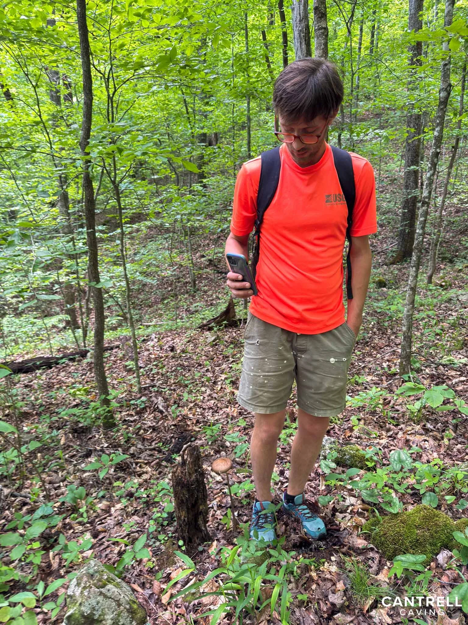 Person in orange shirt and khaki shorts standing in a forest holding a phone, with a backpack and surrounded by trees and greenery.