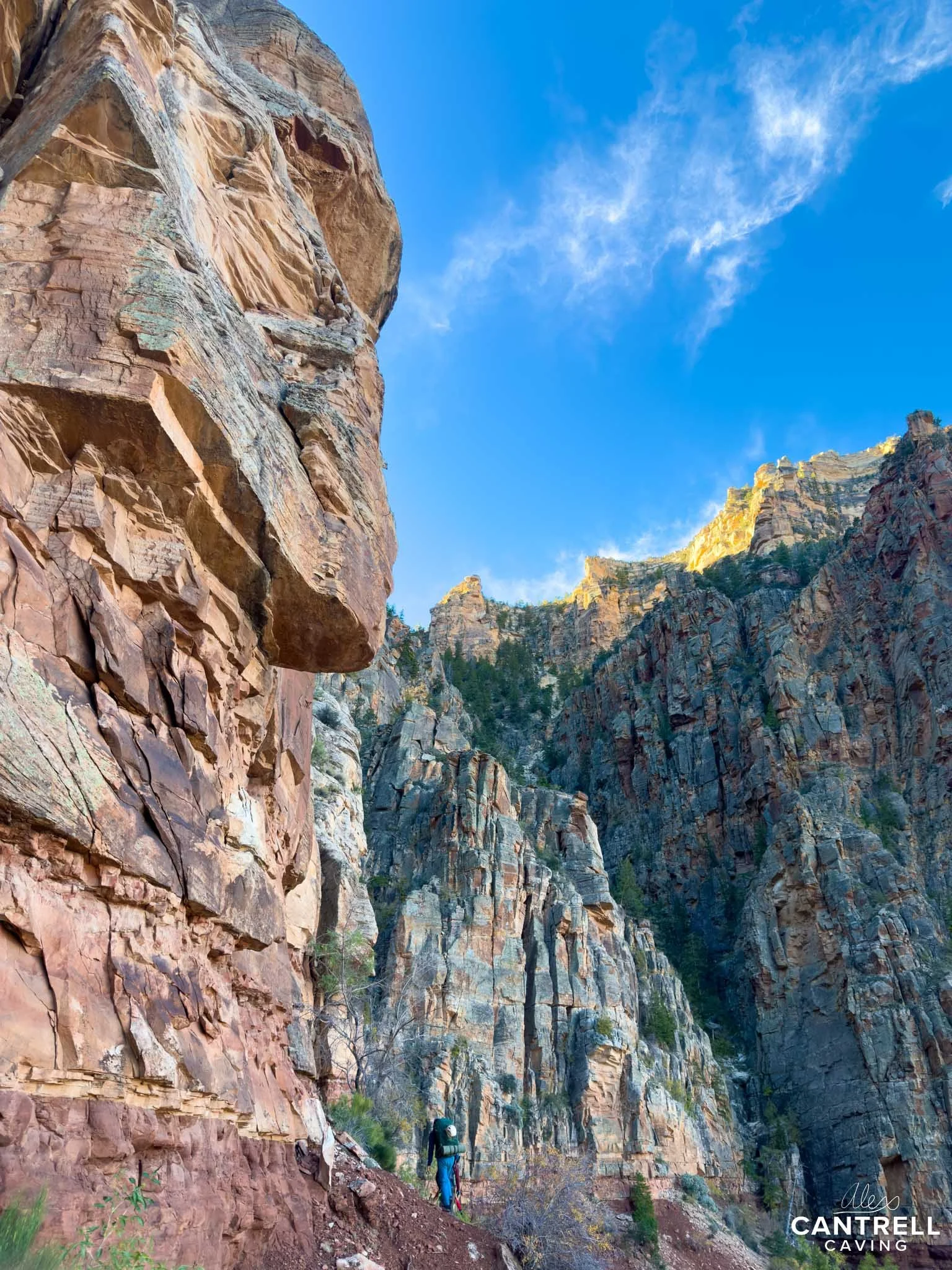 Hiker walking on a trail next to tall rocky cliffs under a clear blue sky.