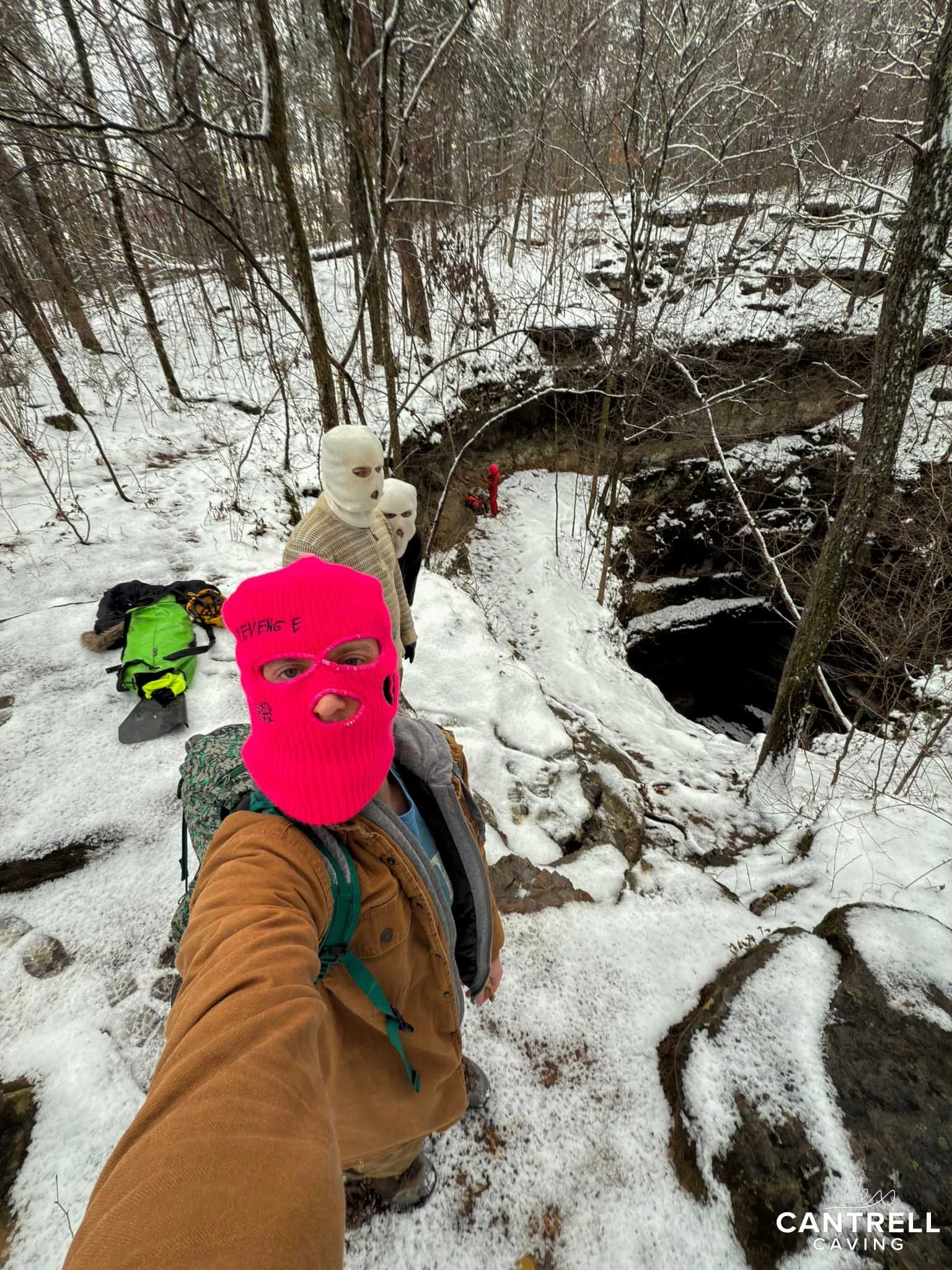 Three people in balaclavas standing near a snowy cave entrance, surrounded by a forest. One person in a pink balaclava is taking a selfie. Snow covers the ground and trees in the background.