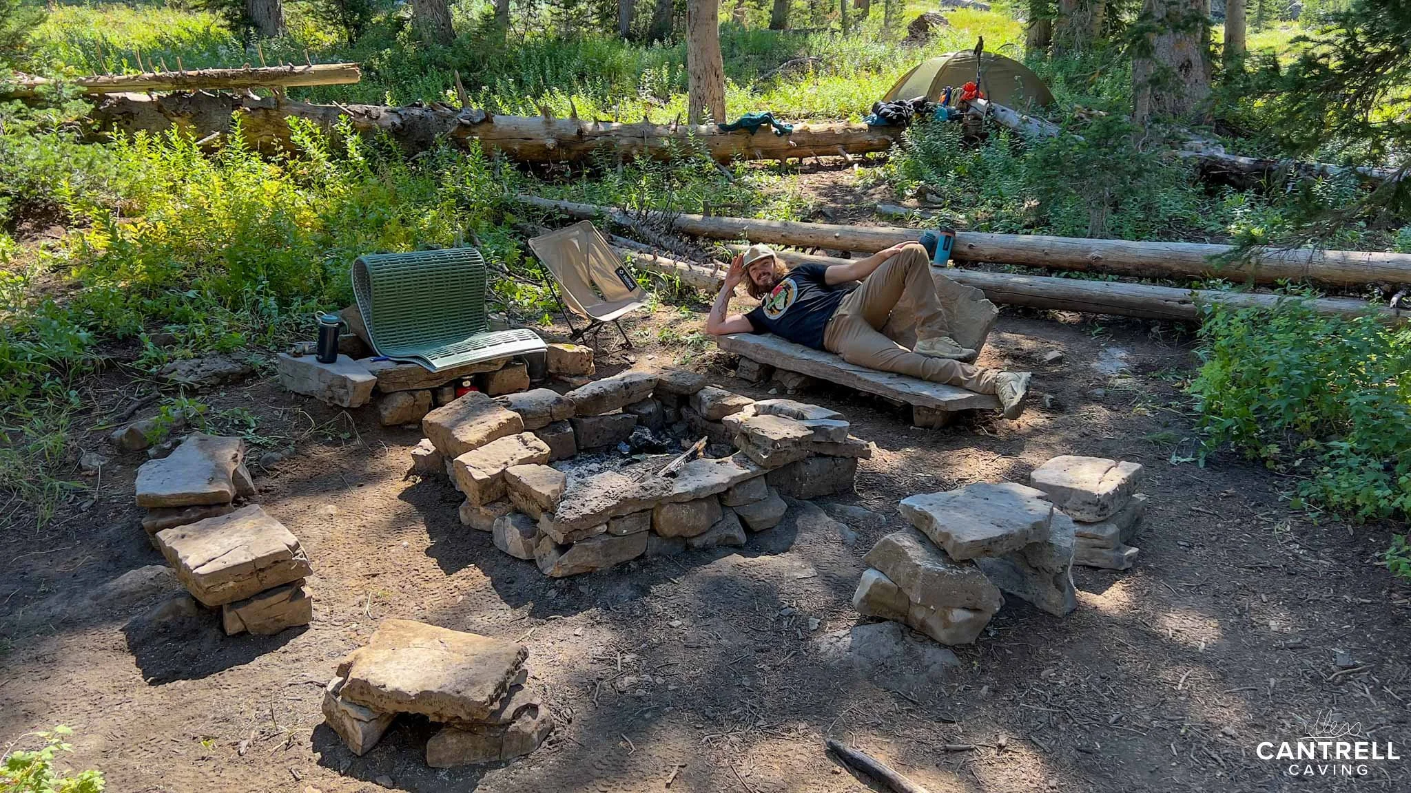 A man relaxing at a rustic campsite in the woods. The setup includes a stone fire pit with surrounding stone seats, a folding chair, and a small table with a cup on it. The man is laying on a makeshift wooden platform. In the background, there is a t