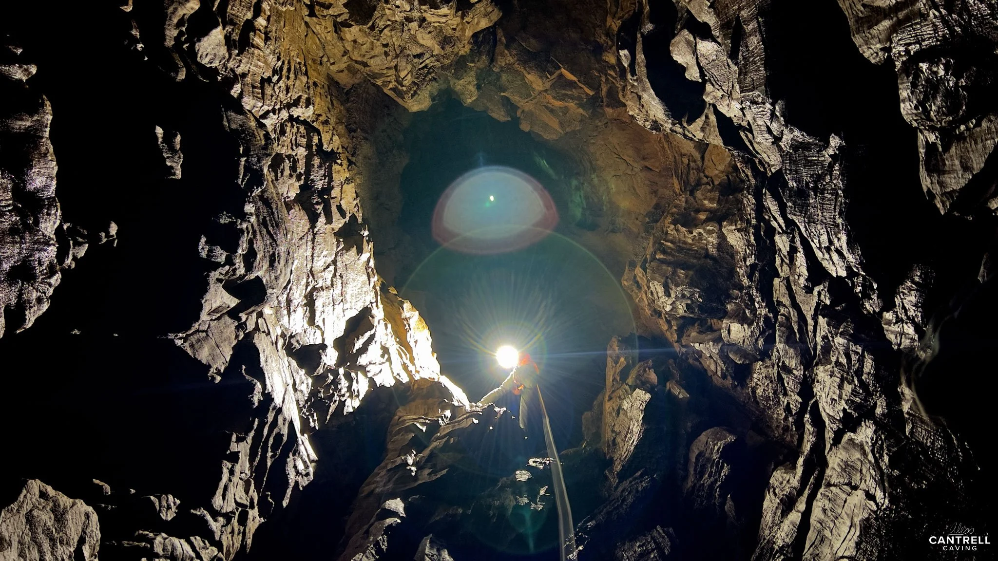 Caver descending into a large rocky cave with bright light shining from the helmet, illuminating the cavern walls.