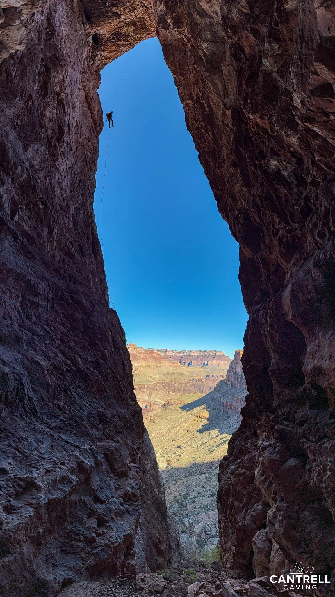 A person rappelling down a rocky canyon with a blue sky above and a landscape visible in the distance.