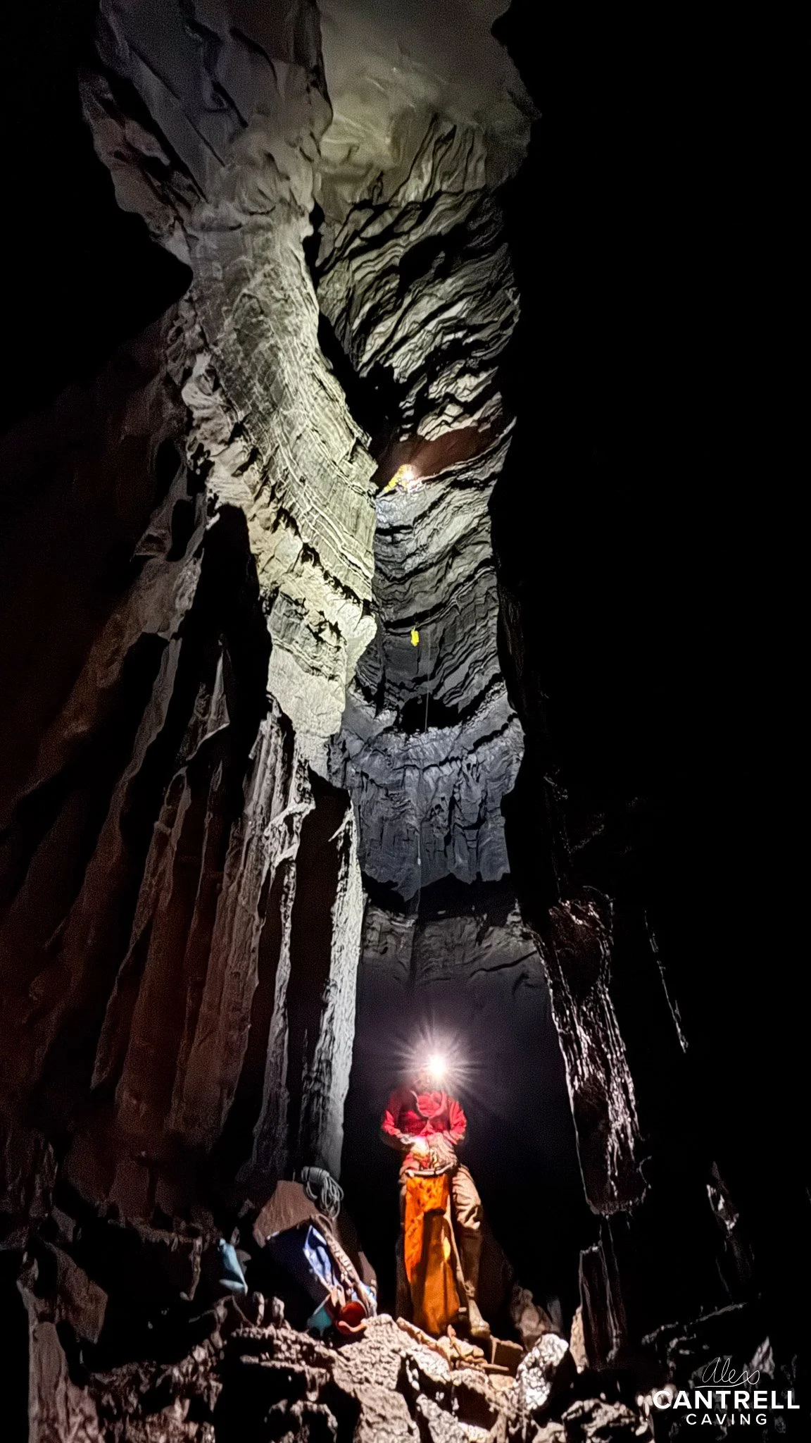 A cave explorer wearing a helmet with a light stands inside a large, dark cavern with steep rock walls. The person is surrounded by caving gear and appears to be preparing for a descent. The rocky interior of the cave is illuminated by the helmet lig