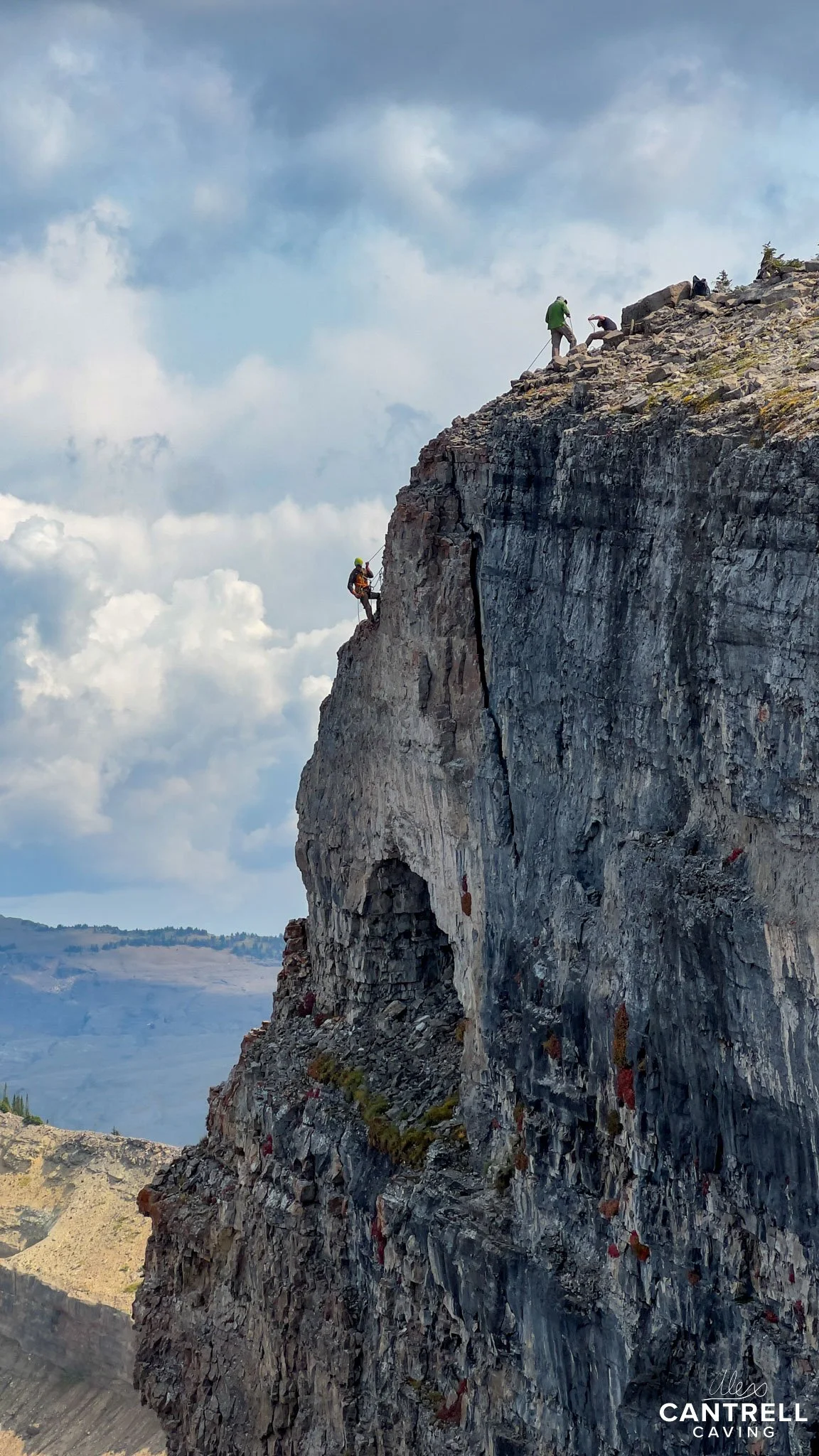 Two rock climbers on a steep cliff, one descending on ropes near the edge, with mountainous landscape in the background.