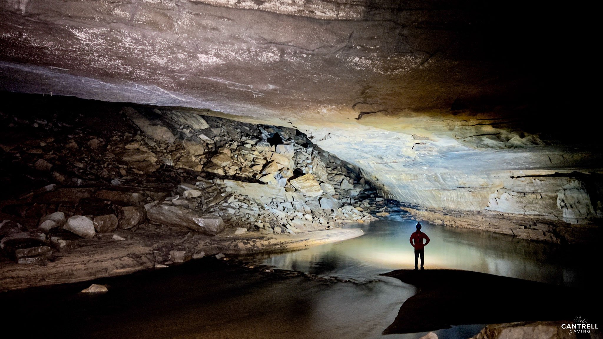 Person standing inside a large cave with rocky walls and a small body of water, illuminated by artificial light.