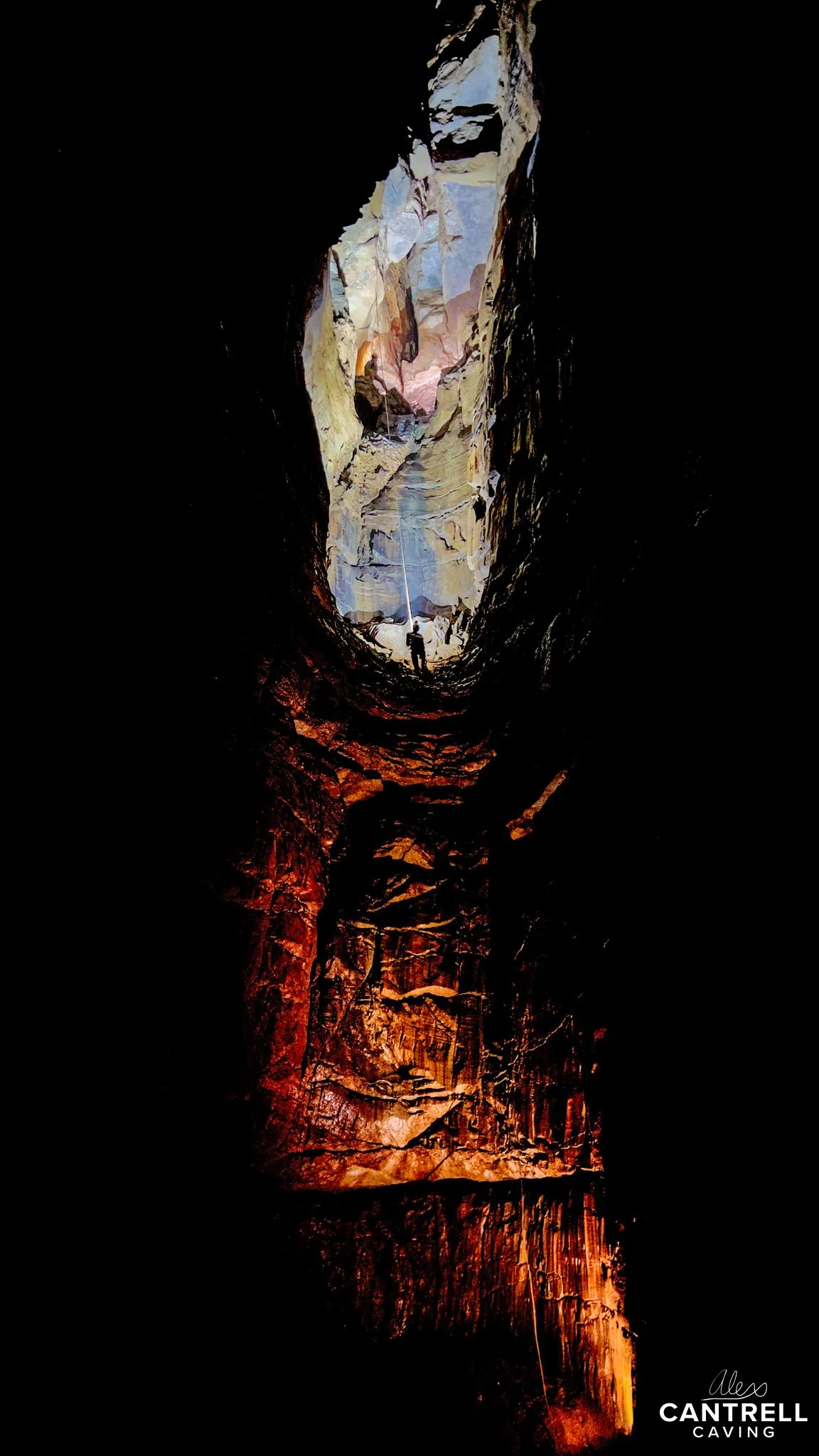 Vertical cave with narrow opening at the top, sunlight illuminating the rocky walls, climber visible near the entrance, deep shadows below. Credit: Alex Cantrell Caving.