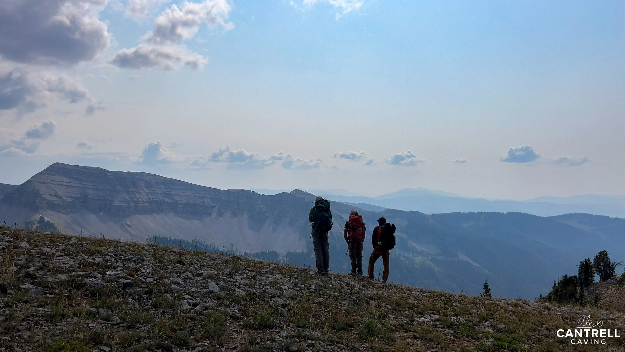 Three hikers with backpacks standing on a rocky hilltop with mountains and cloudy sky in the background.