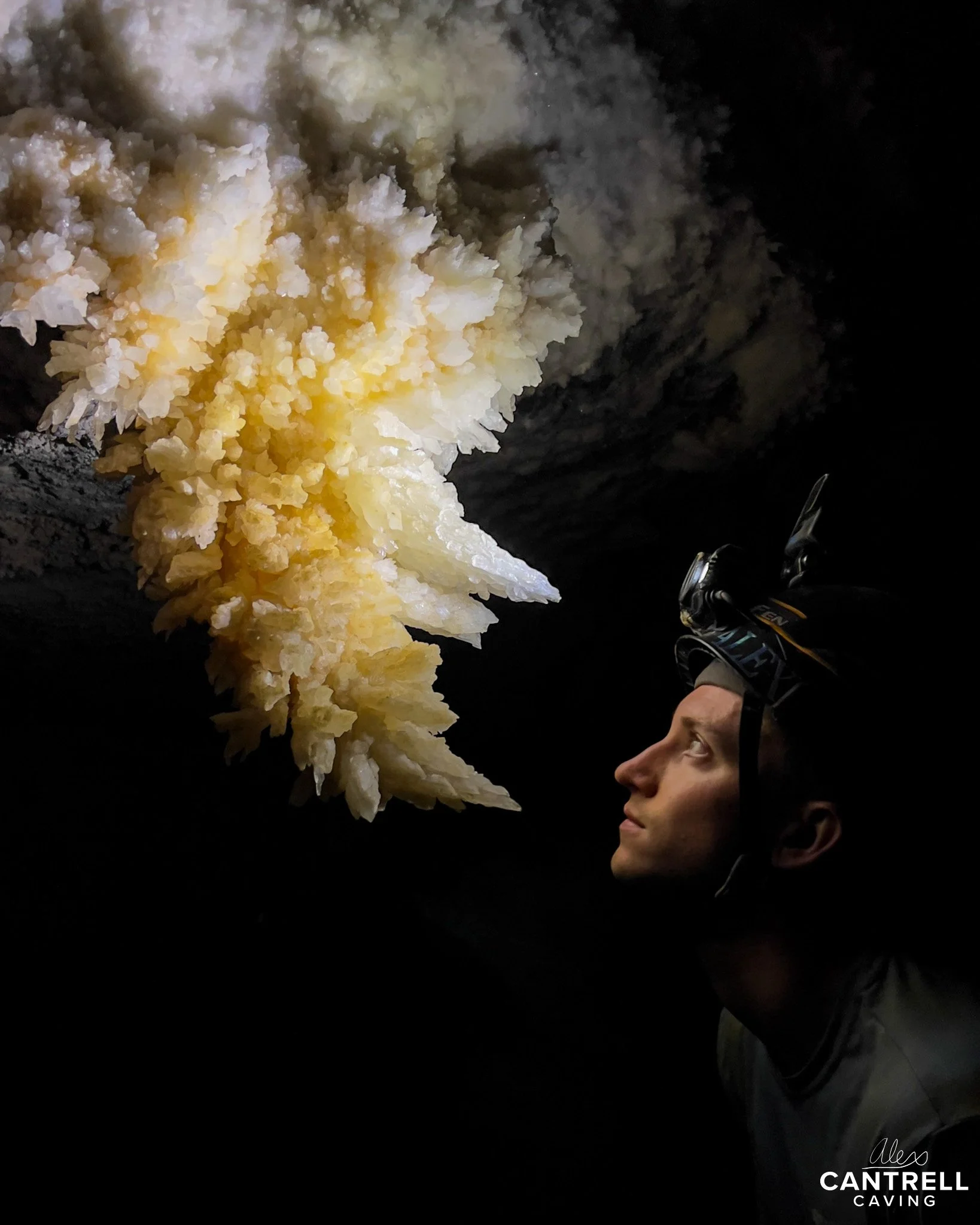 A person wearing a helmet looks at white and yellow crystalline formations on a cave ceiling.
