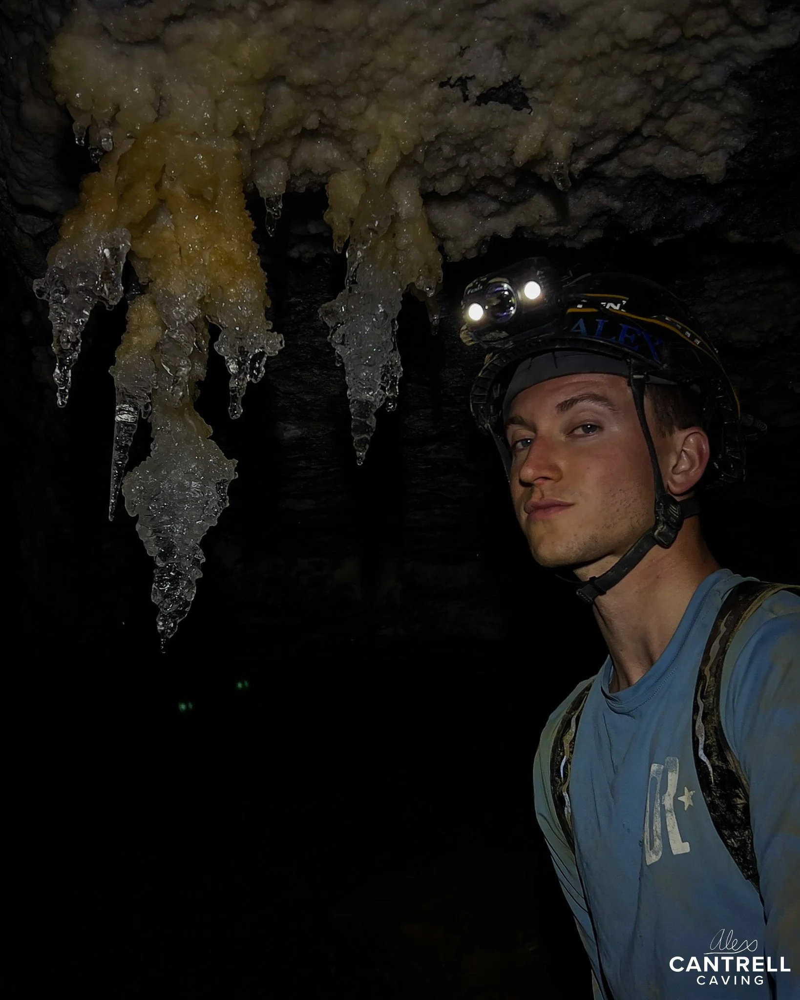 Man wearing helmet with headlamp inside a cave, standing near stalactites on the ceiling.
