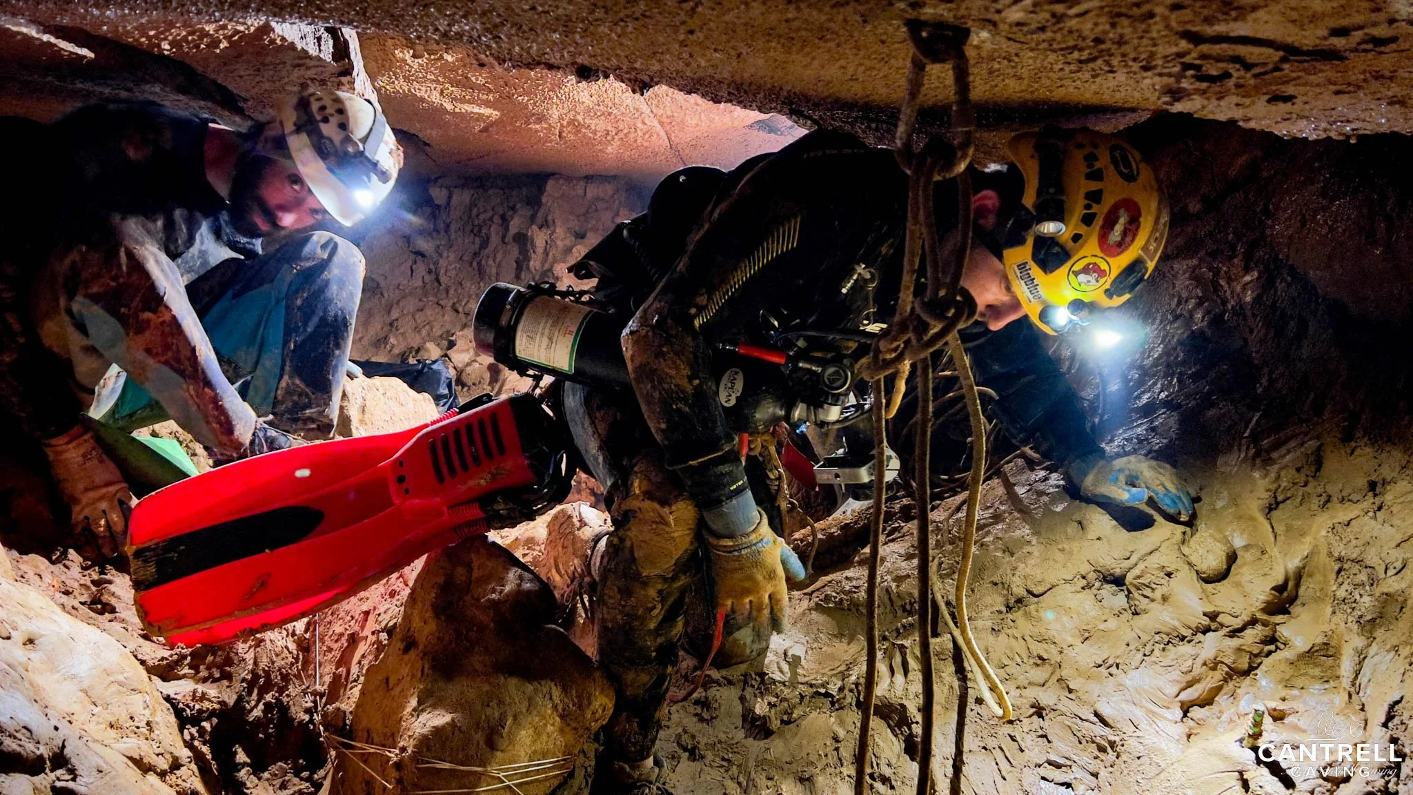 Cave divers navigating through a narrow, muddy passage with headlights and diving equipment.
