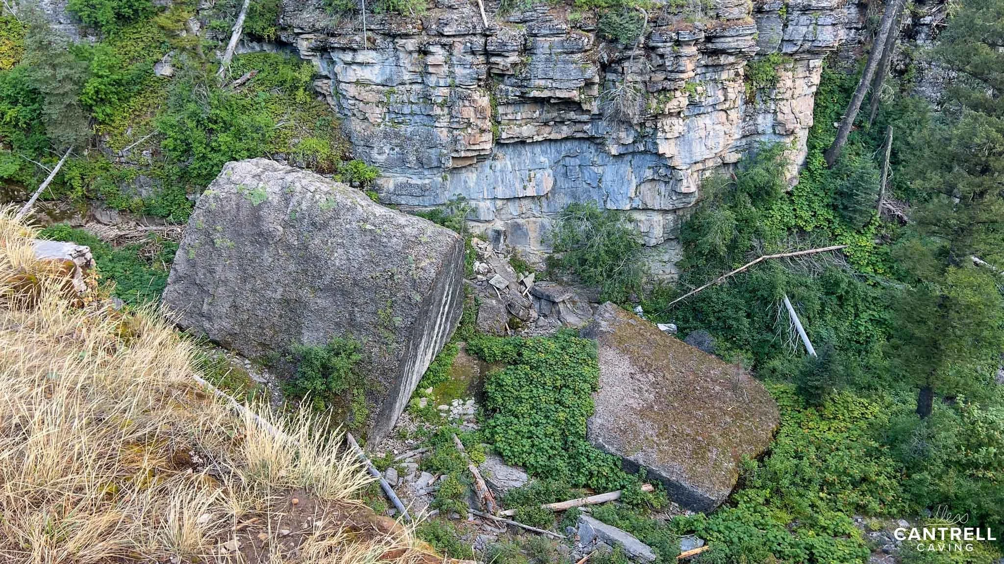Large boulders at the base of a rocky cliff surrounded by lush vegetation and trees.