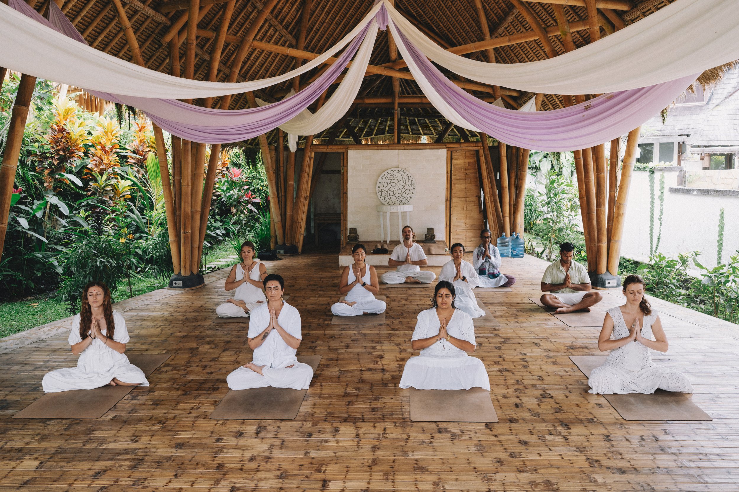 Yoga Teacher Training students in Bali seated in white attire with folded hands and closed eyes during meditation practice