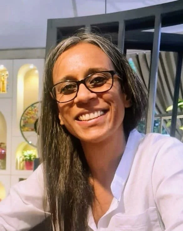 A woman with long dark hair, glasses, and a white shirt smiling at the camera inside a store or cafe with shelves and decorations in the background.