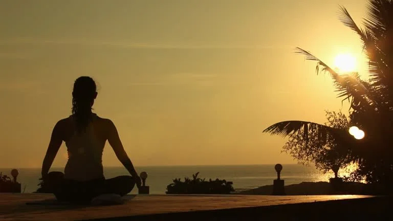 Person sitting in a meditative pose on the beach at sunset, overlooking the ocean in Bali