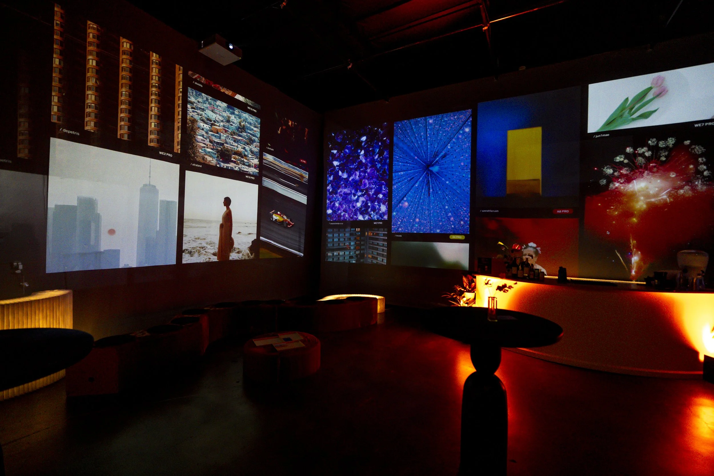 Dark room with multiple screens displaying colorful images including cityscapes, abstract patterns, and fireworks. The room has a soft warm glow from a few lamps and appears to be a modern lounge or gallery.