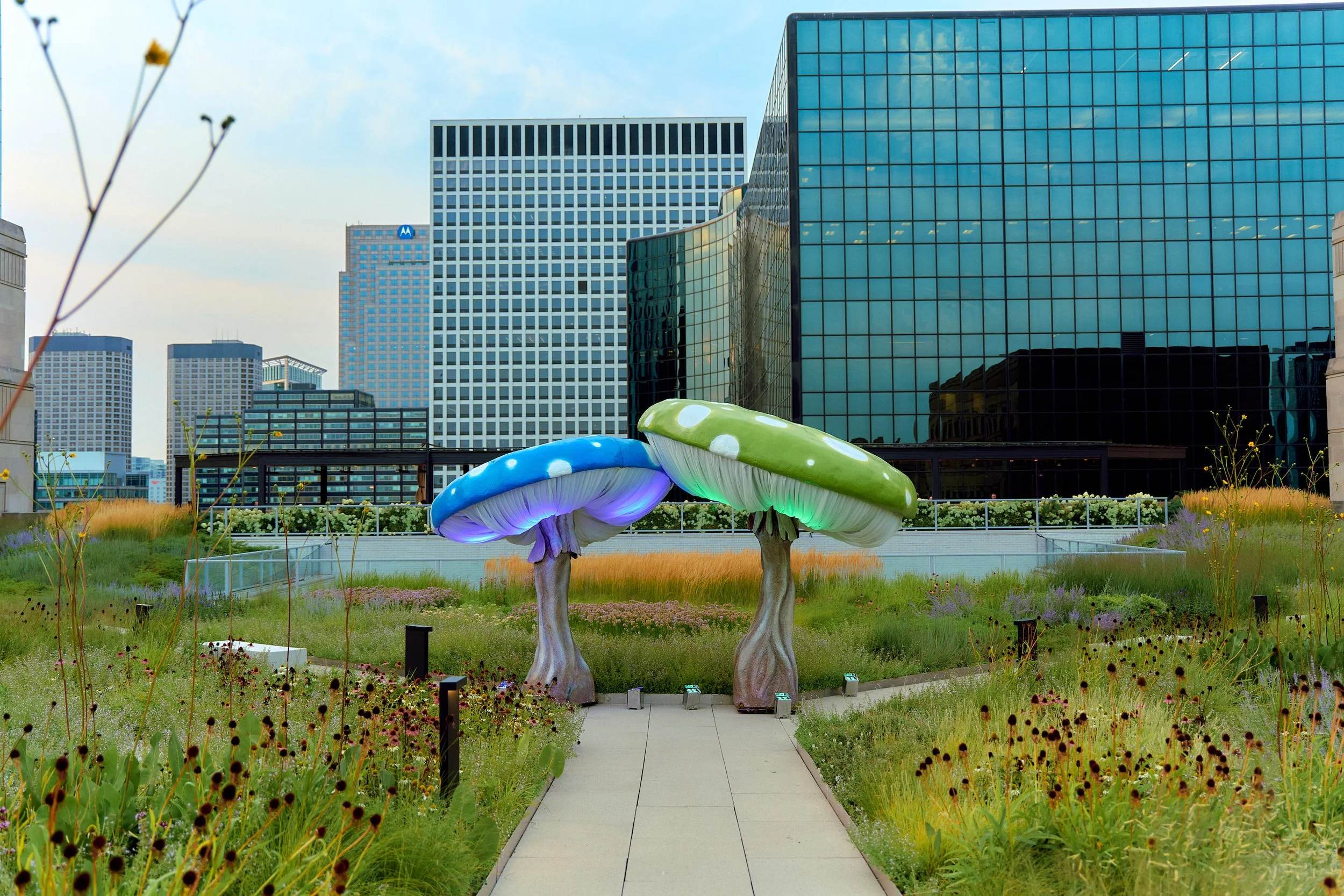 Two large, colorful mushroom sculptures, one blue and one green, stand in a landscaped rooftop garden in an urban cityscape with tall modern buildings in the background.