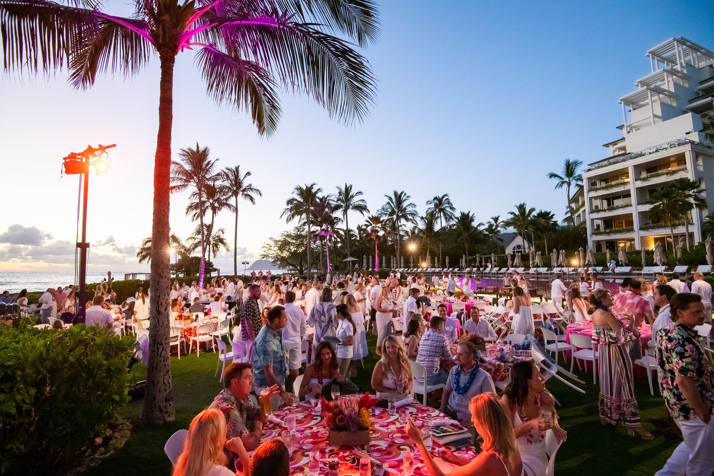 Outdoor tropical resort party at sunset with palm trees, people in casual summer clothes, tables with pink and red decorations, and white building balconies in the background.