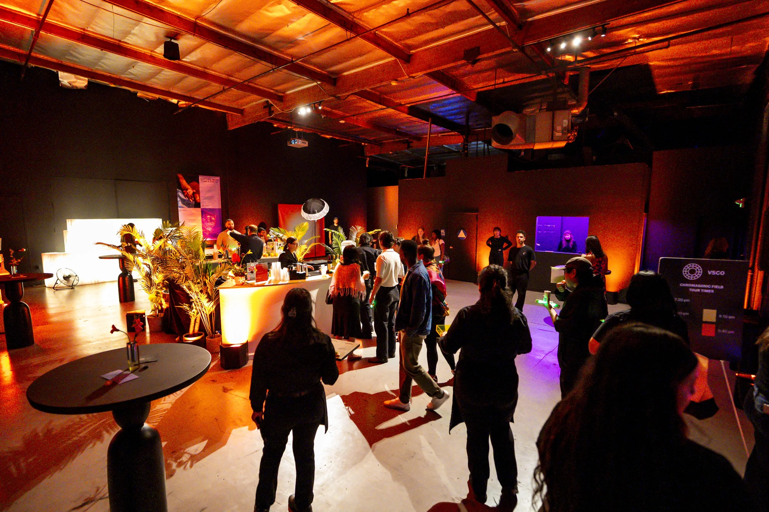 People attending an indoor event or social gathering in a dimly lit room with colorful lighting, a bar area, and several booths or displays.