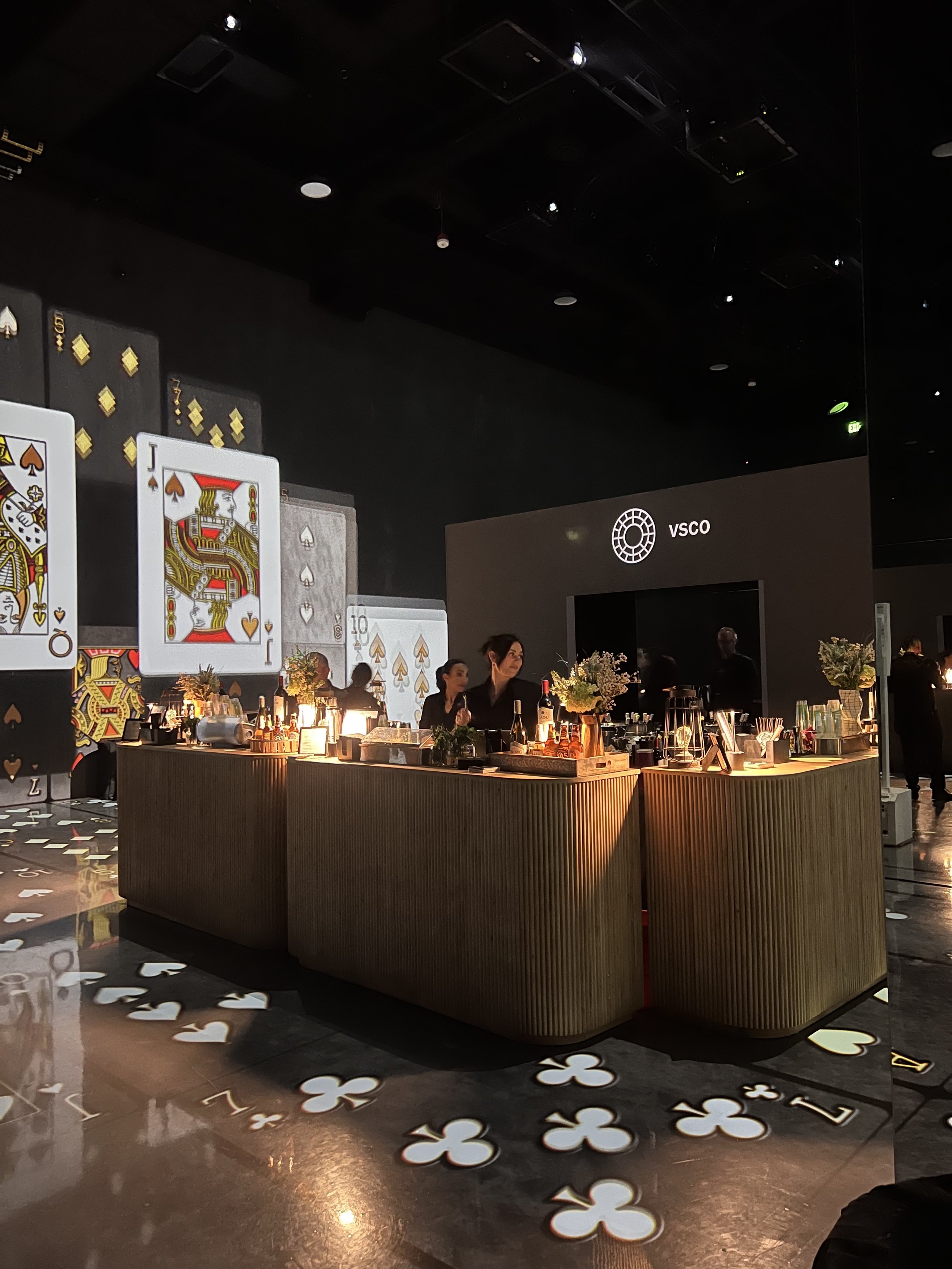 A bar with a dark background, decorated with large playing cards and illuminated by warm lighting. There are bartenders behind the counter, and the VSCO logo displayed on a screen in the background.