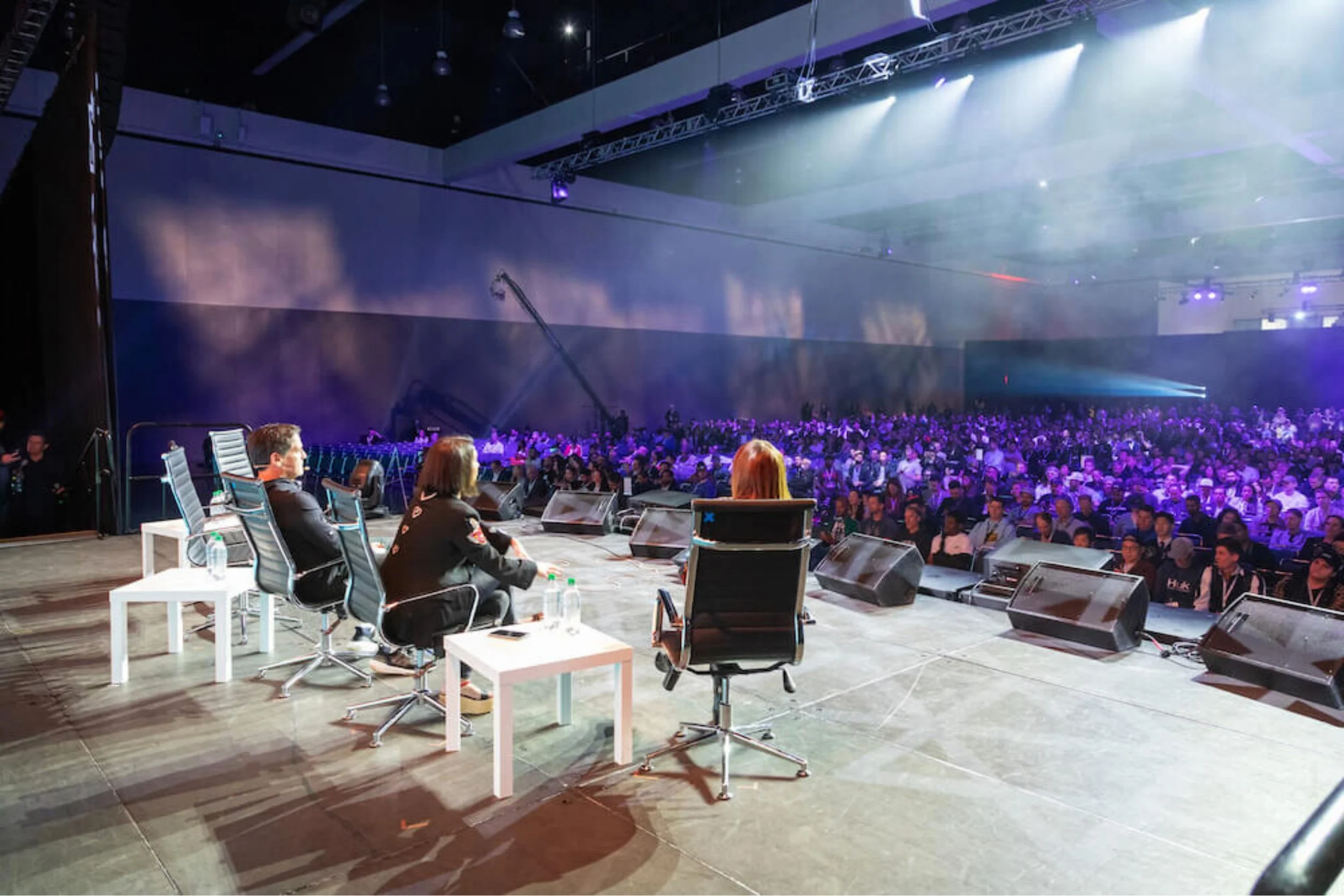 Three individuals on stage facing an audience in a large conference hall with purple and white lighting, speakers, and screens.