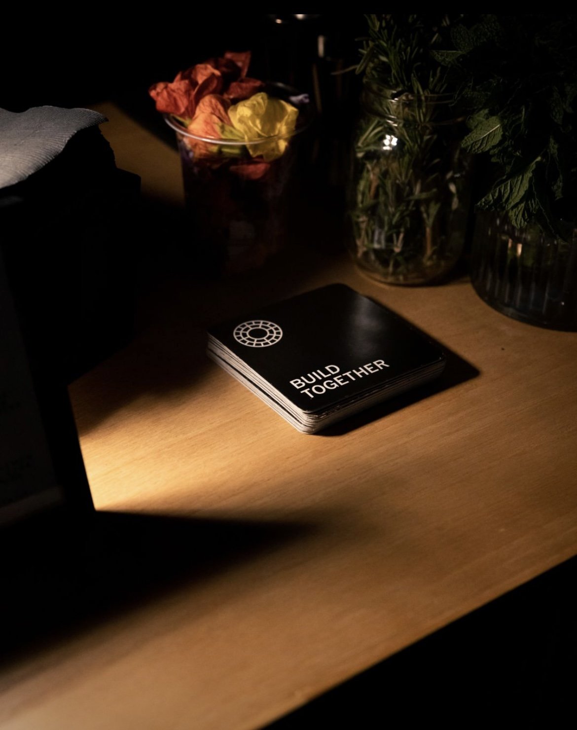 A deck of black cards with the words 'BUILD TOGETHER' and a wheel symbol on top, placed on a wooden surface illuminated by a focused light.