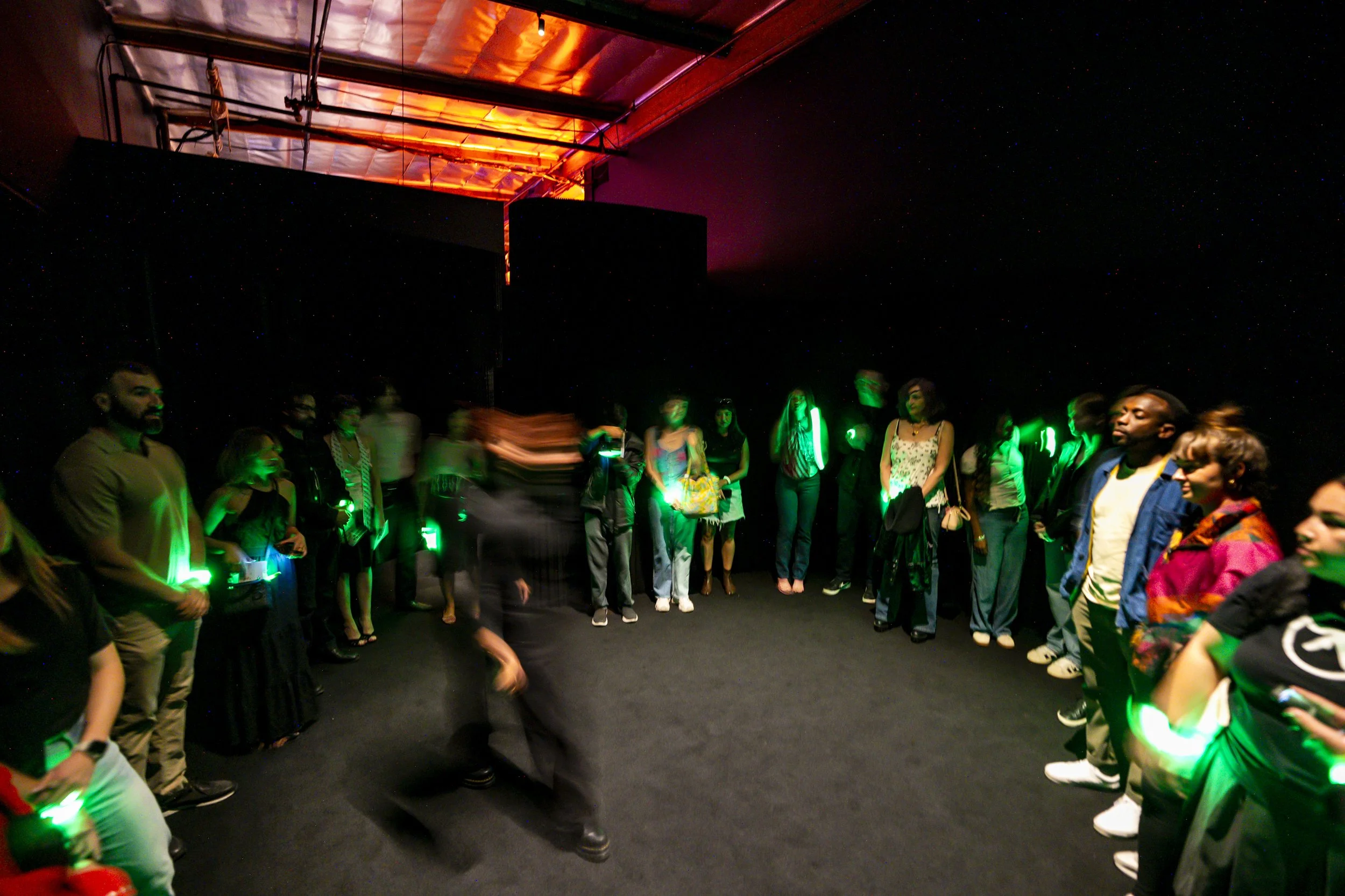Group of people standing in a circle in a dark room with green glow sticks. There is a person walking across the circle, creating a blur. The room has a black wall and a ceiling with industrial pipes and a reflective surface.