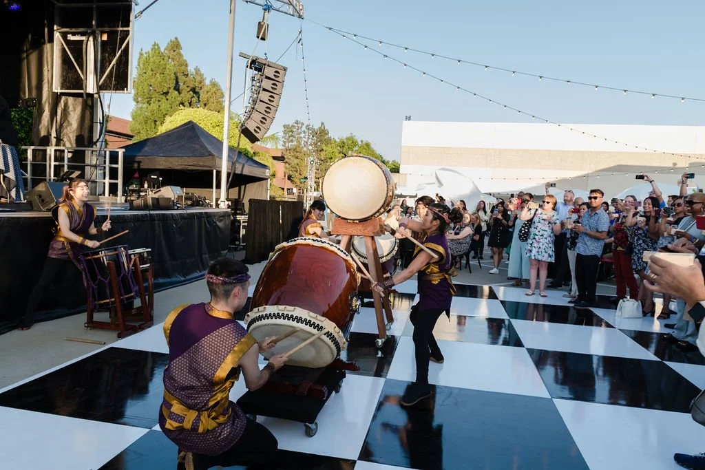 Performers playing traditional drums at an outdoor event with a black-and-white checkered dance floor, a stage, and an audience watching and taking photos.