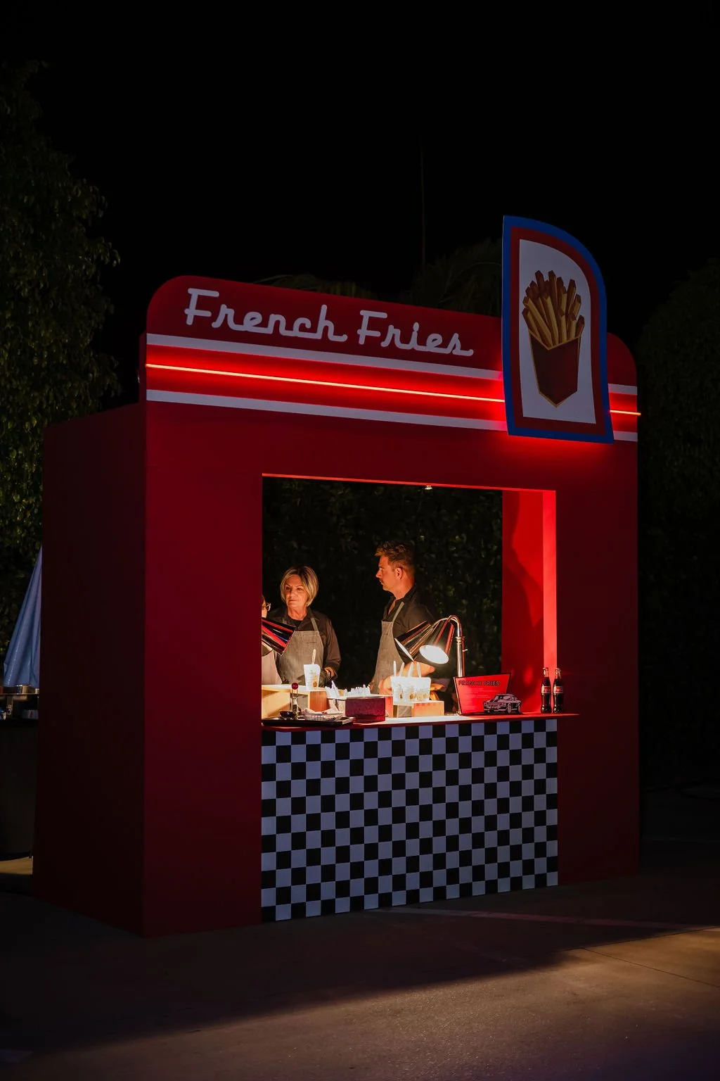 Nighttime outdoor food stand with a red and white striped sign that reads 'French Fries' and a picture of French fries. Two people wearing aprons are working inside the stand, which has a black and white checkered pattern at the front.