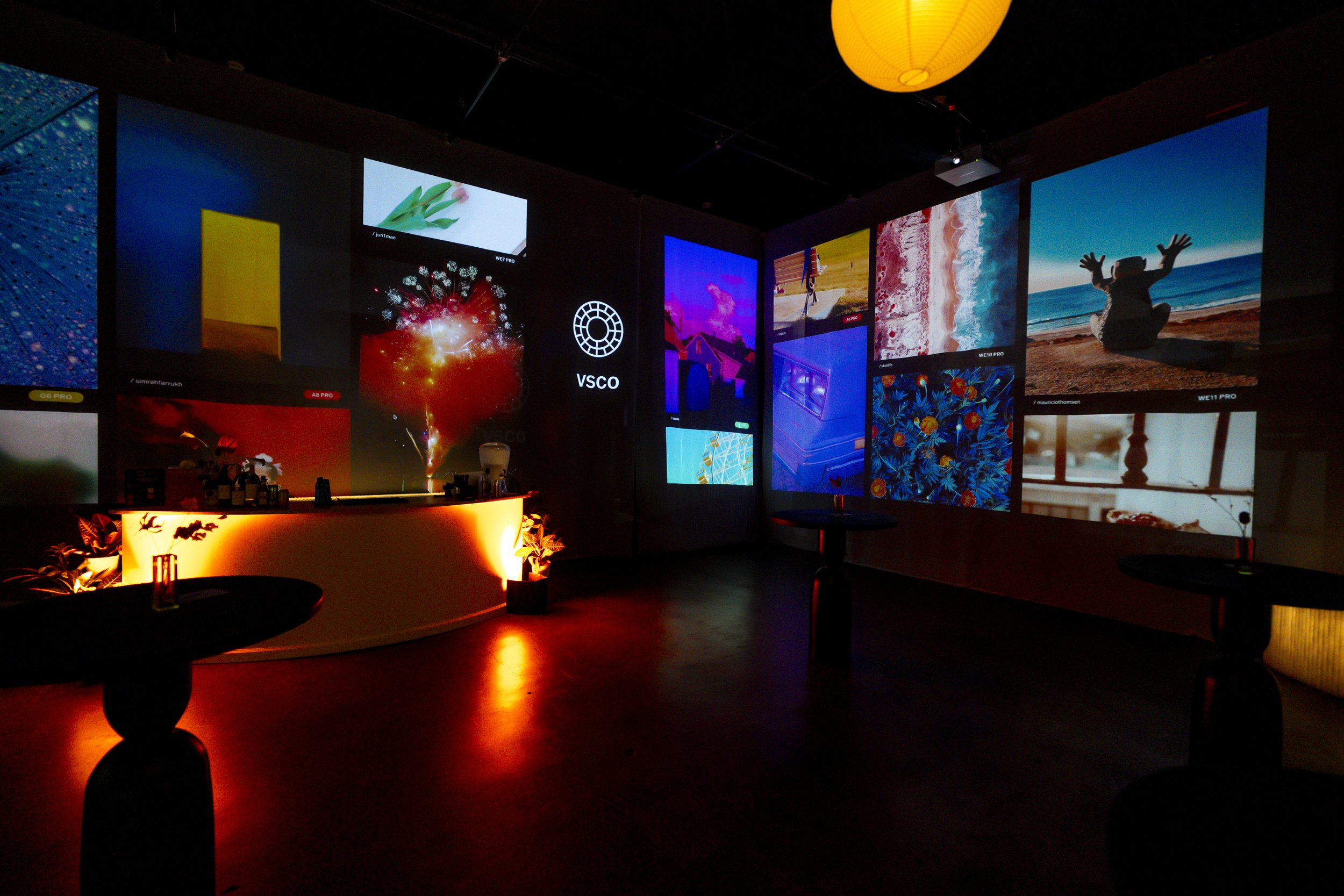 Dark room with a curved lighted bar, multiple large colorful screens displaying various images, some tables and plants, and a yellow paper lantern hanging from the ceiling.