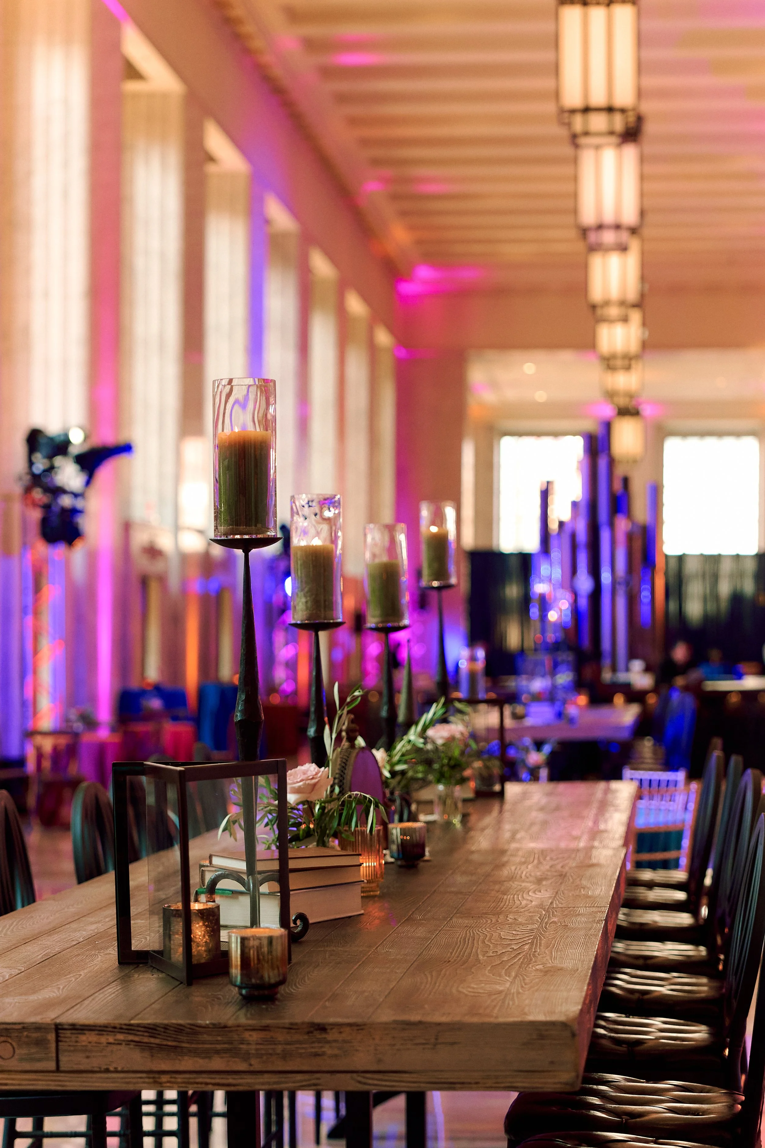 A decorated event space with a long wooden table, tall candles in glass holders, floral arrangements, and candles, with colorful ambient lighting and large windows in the background.