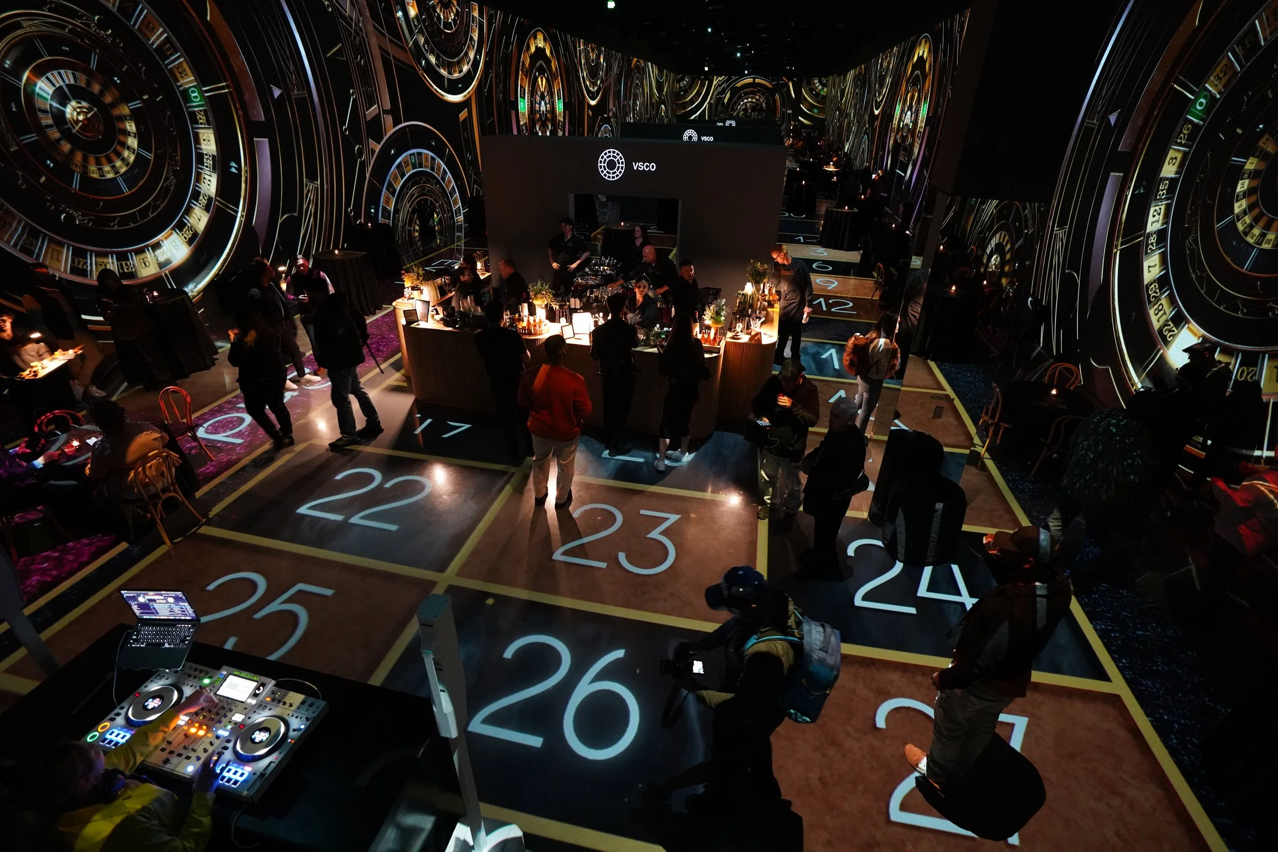 A lively indoor event space with many people gathered around tables and a central bar area. The room features large digital roulette wheel graphics on the walls and floor, with numbered sections and neon lighting. A DJ setup is visible in the foreground, and the atmosphere appears energetic and modern.